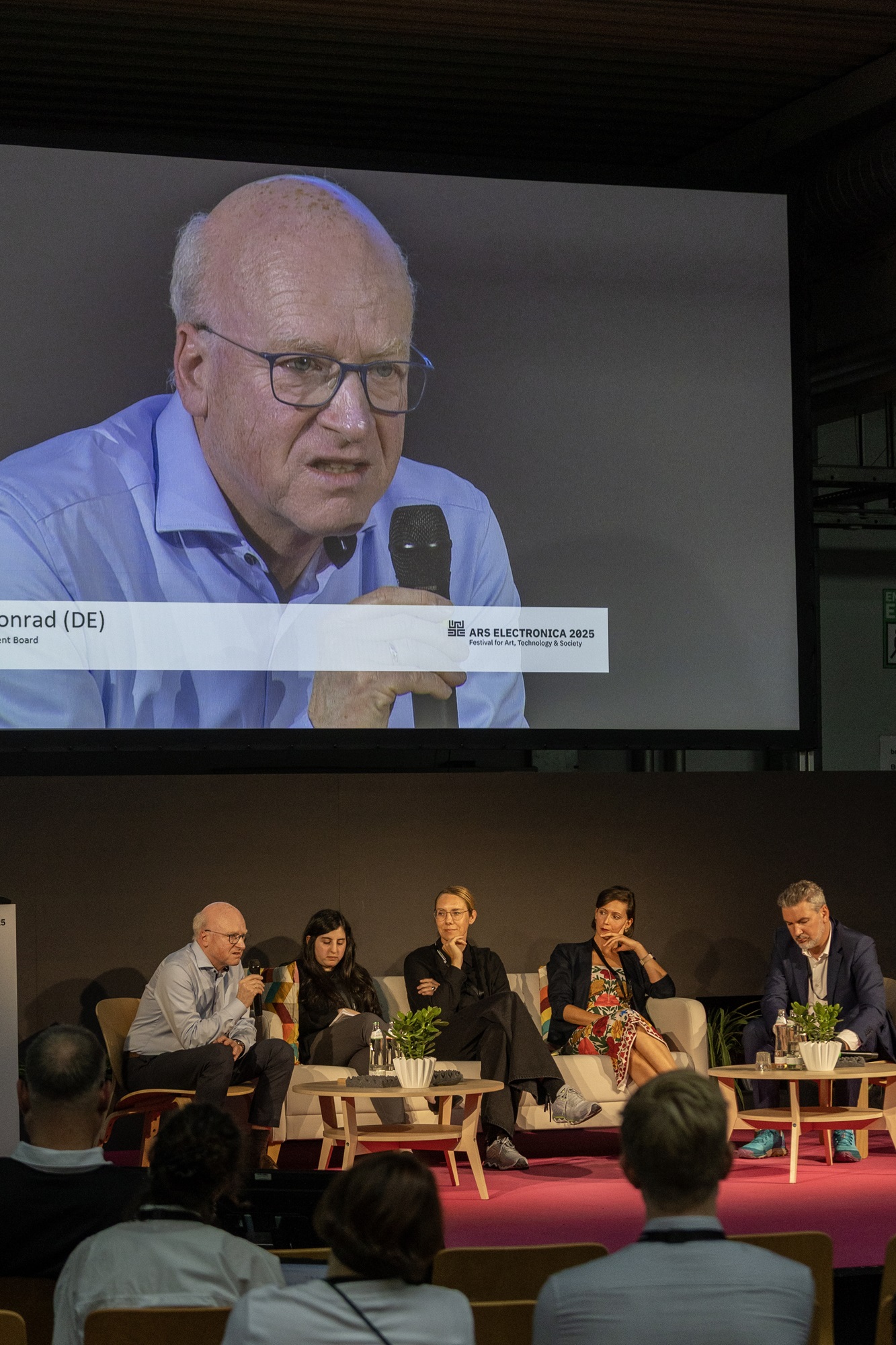 Auf einer Bühne sitzen fünf Personen nebeneinander in Sesseln. Matthias Konrad, Mitglied der Geschäftsleitung bei der Bayern Innovativ GmbH, spricht ins Mikrofon. Über den Köpfen hängt eine große Leinwand, auf der Matthias Konrad in Nahaufnahme zu sehen ist. Vor der Bühne sitzt Publikum.