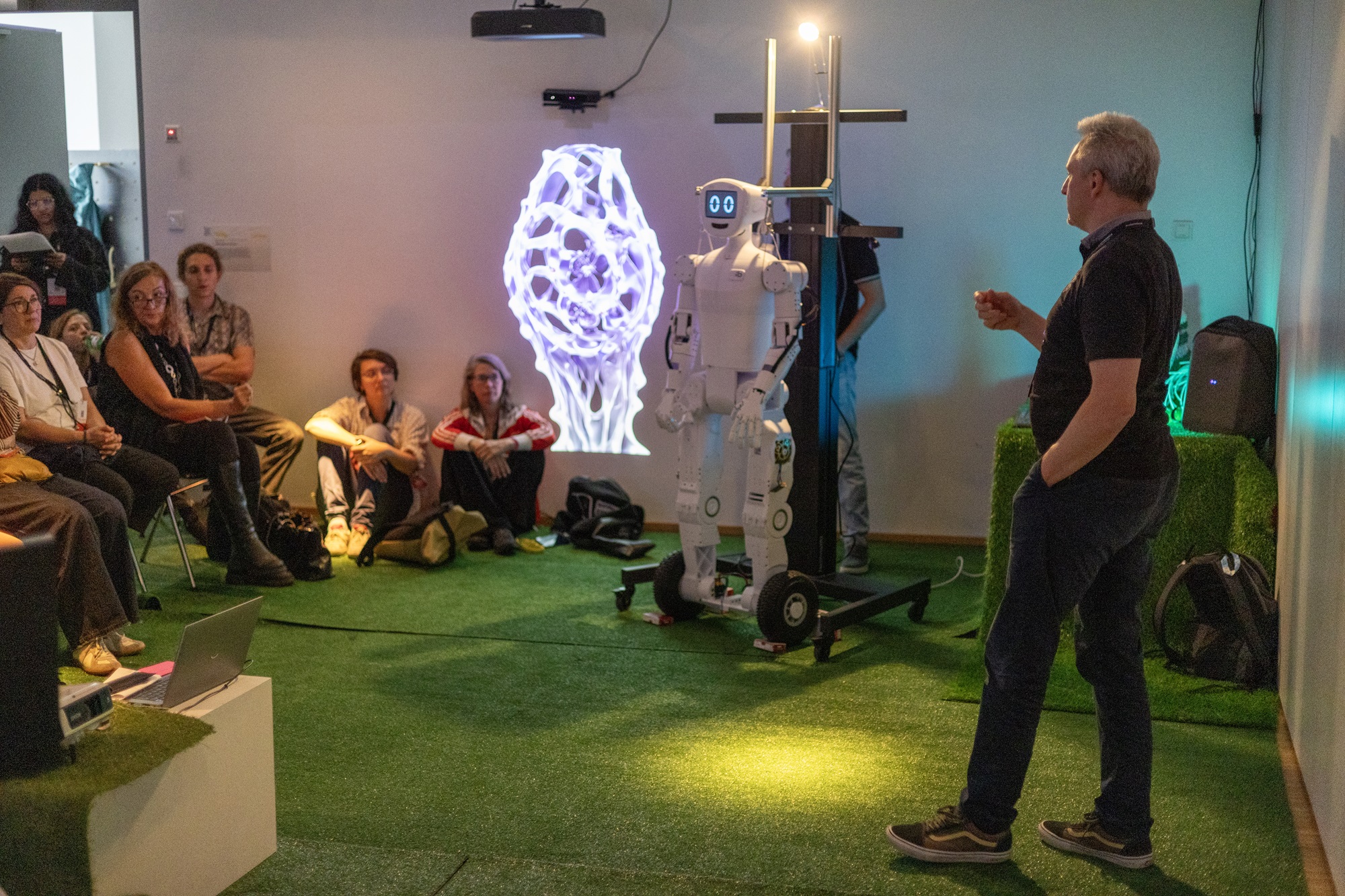 In einem Raum mit grünem Teppichboden steht Andreas Böhm, Innovationsnetzwerk Digitalisierung, Leitung ThinkNet Quantentechnologie, Bayern Innovativ GmbH und spricht zur Gruppe. Neben ihm steht ein weißer Roboter auf Rädern. Rundherum sitzen Menschen auf Stühlen und am Boden und hören zu. An der Wand hinter dem Roboter leuchtet eine große Projektion in hellen Farben.