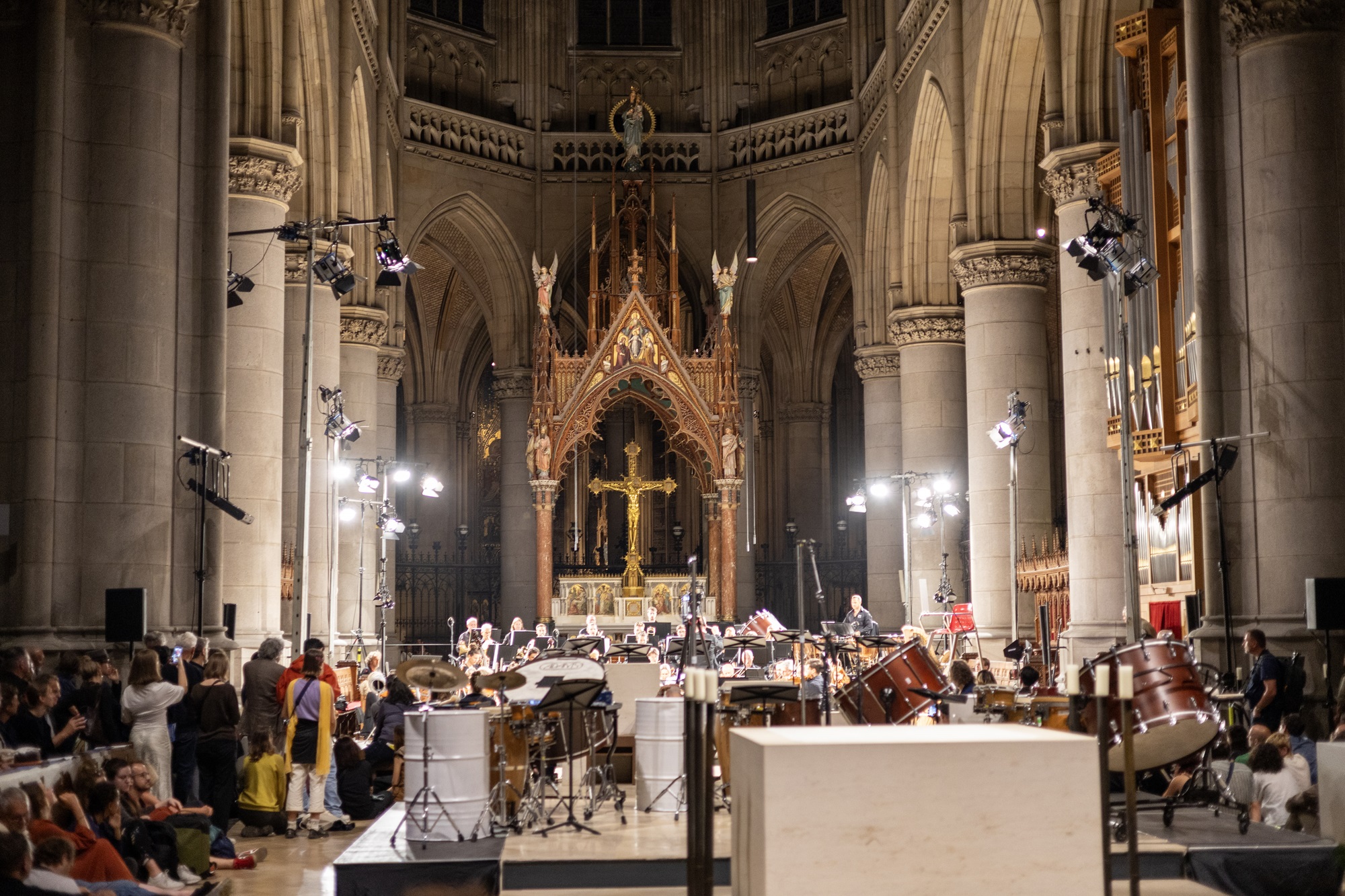 In einer großen Kirche sitzen viele Menschen im Publikum und hören einem Orchester zu. Im Vordergrund steht ein Schlagzeug mit weiteren Instrumenten. Im Hintergrund spielt das Orchester direkt vor dem Altar. In der Mitte des Altars ist ein großes goldenes Kreuz. Helle Scheinwerfer erleuchten den ganzen Raum.