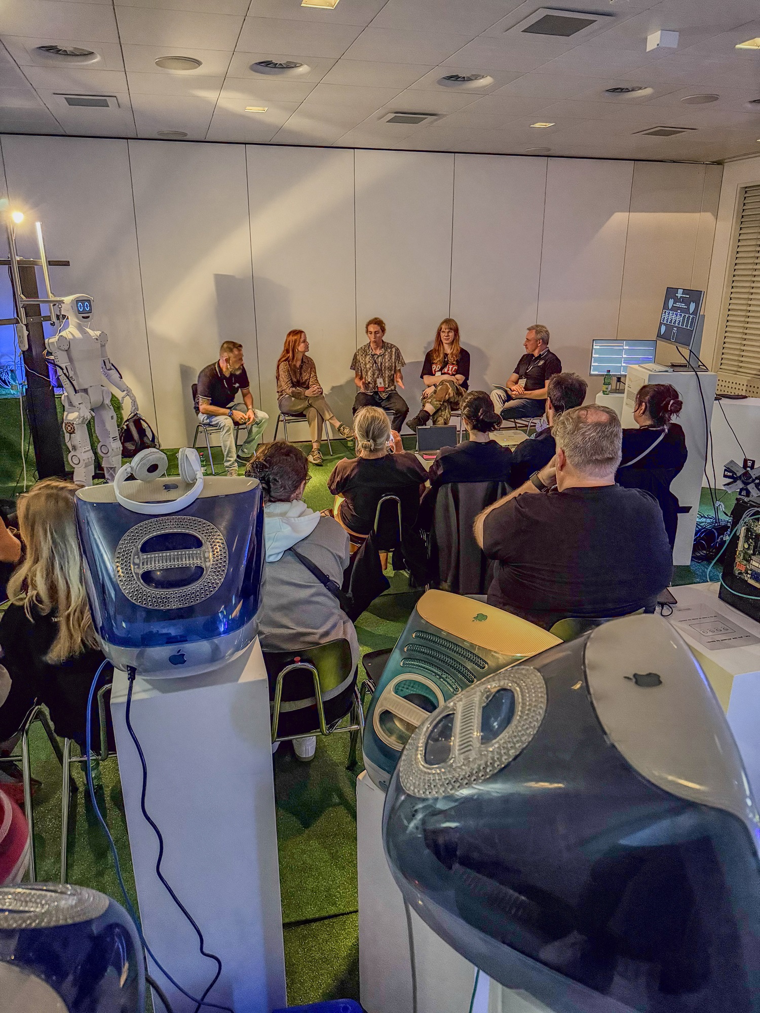 In einem Raum mit grünem Boden sitzt ein Publikum und schaut nach vorne. Vorne sitzen fünf Personen nebeneinander in einer Gesprächsrunde, darunter Andreas Böhm, Innovationsnetzwerk Digitalisierung, Leitung ThinkNet Quantentechnologie, Bayern Innovativ GmbH. Rechts hinten im Raum steht ein Bildschirm mit technischen Anzeigen. Links am Rand steht ein weißer Roboter. Im Vordergrund sind mehrere bunte Computergehäuse zu sehen.