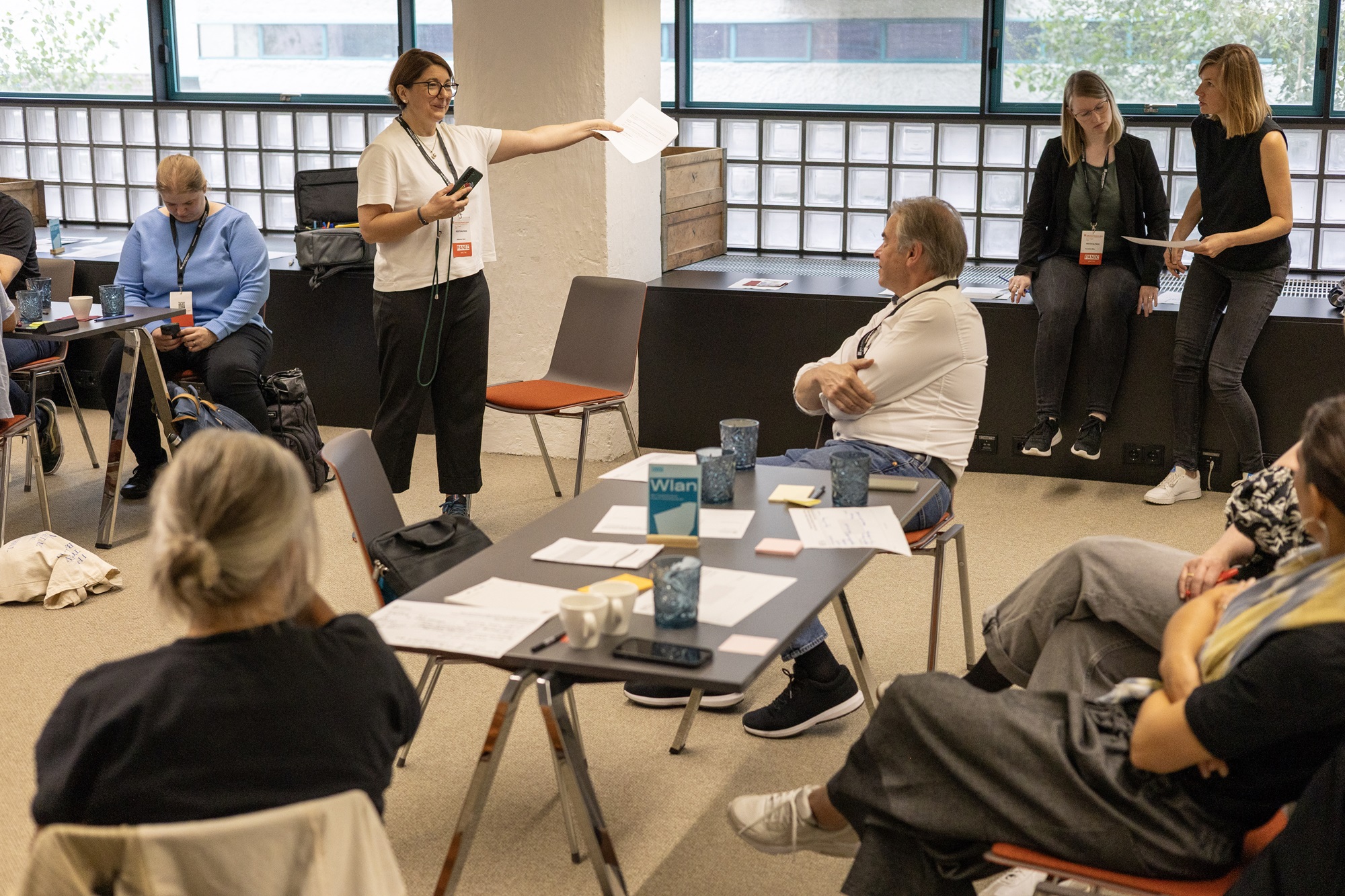 In einem Raum sitzen mehrere Menschen an Tischen, auf denen Blätter und Gläser liegen. Vorne steht Johanna Lison, Projekt- und Innovationsmanagerin im Innovationsnetzwerk Kreativwirtschaft der Bayern Innovativ GmbH. Sie spricht zur Gruppe und zeigt mit einem Blatt Papier nach vorn. Einige Personen hören aufmerksam zu, während andere am Rand des Raumes sitzen und beobachten.