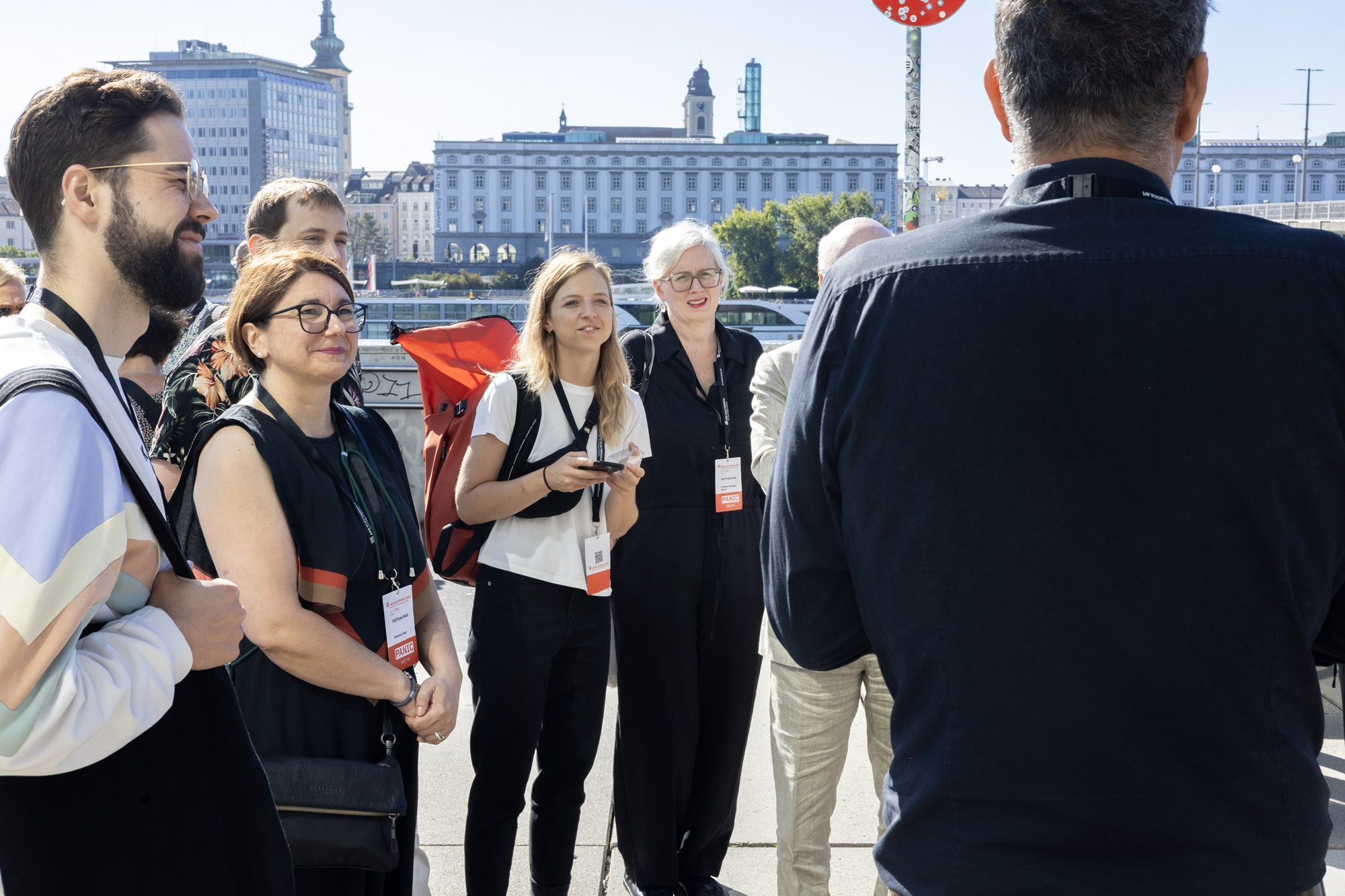 Eine Gruppe von Menschen steht draußen im Sonnenschein und hört einer Person zu, die einen Vortrag hält. Unter den Teilnehmenden sind Johanna Lison, Projekt- und Innovationsmanagerin im Innovationsnetzwerk Kreativwirtschaft bei der Bayern Innovativ GmbH, sowie Julia Wirnshofer, Projektmanagerin im Innovationsnetzwerk Kreativwirtschaft bei der Bayern Innovativ GmbH. Im Hintergrund sind die Gebäude der Stadt Linz zu sehen.