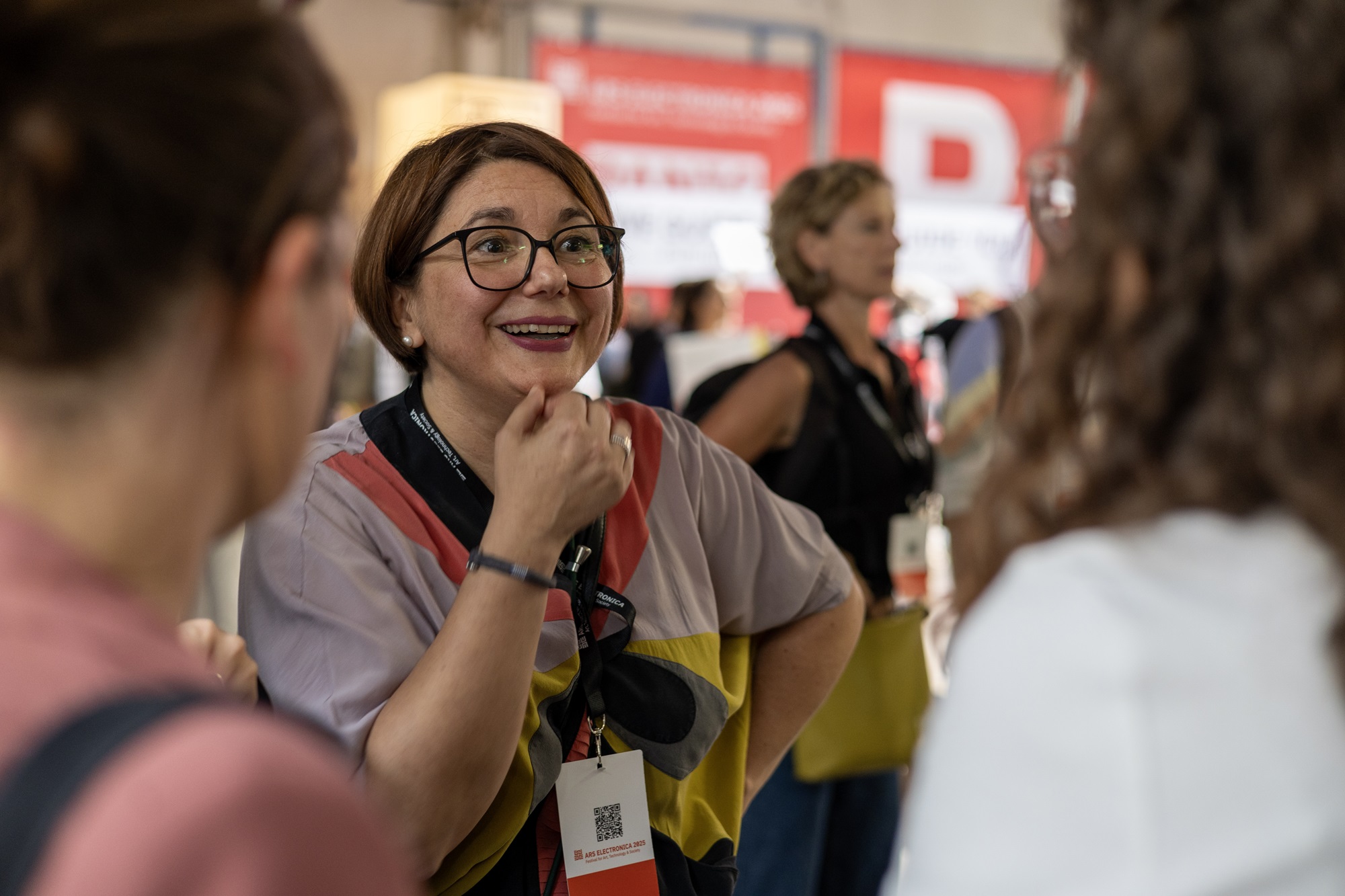 Johanna Lison, Projekt- und Innovationsmanagerin im Innovationsnetzwerk Kreativwirtschaft der Bayern Innovativ GmbH, unterhält sich lächelnd mit anderen Personen. Im Hintergrund sind weitere Menschen und rote Banner einer Veranstaltung zu erkennen.