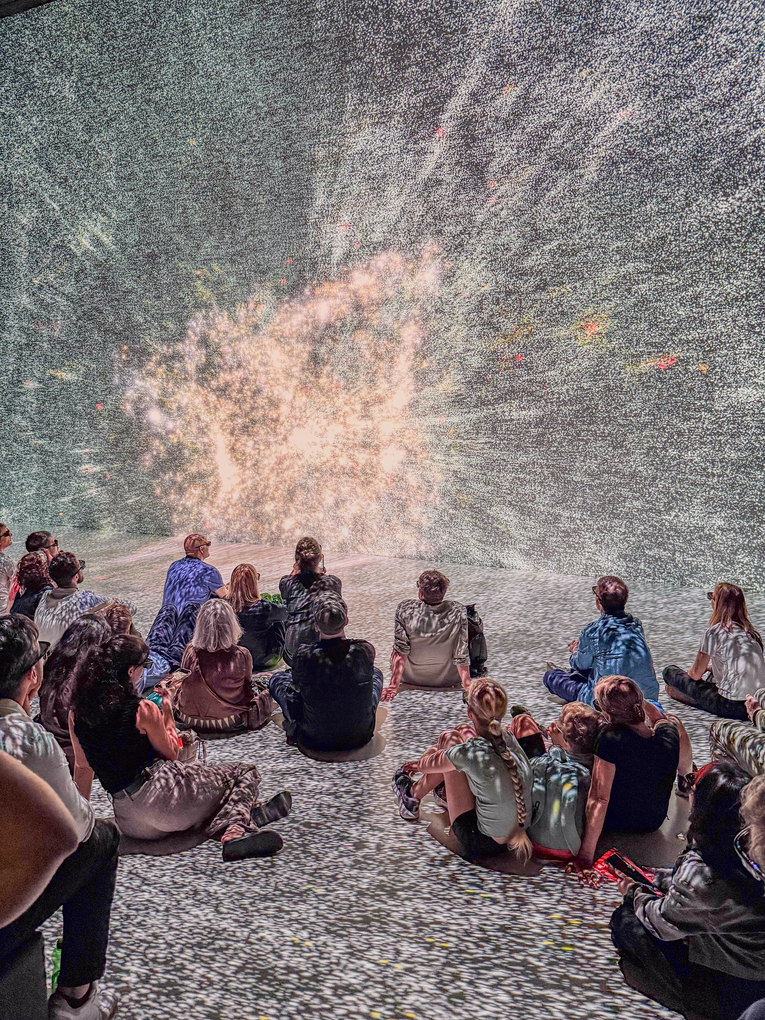 Viele Menschen sitzen auf dem Boden und schau-en nach vorn. An den Wänden und auf dem Boden ist eine große Projektion zu sehen. In der Mitte leuchtet ein heller Punkt und von dort strahlen viele kleine Lichtpunkte nach außen. 