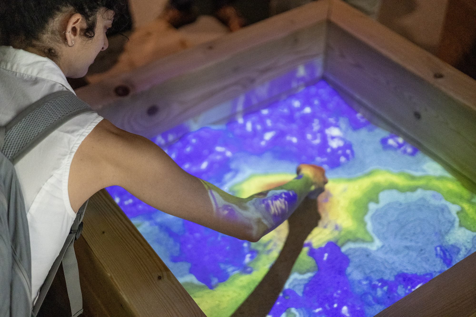 Links im Bild ist eine Person zu sehen, die mit der Hand in einen interaktiven Sandkasten greift. Auf die Sandfläche wird eine farbige Projektion geworfen, die wie eine Landschaftskarte aussieht.
