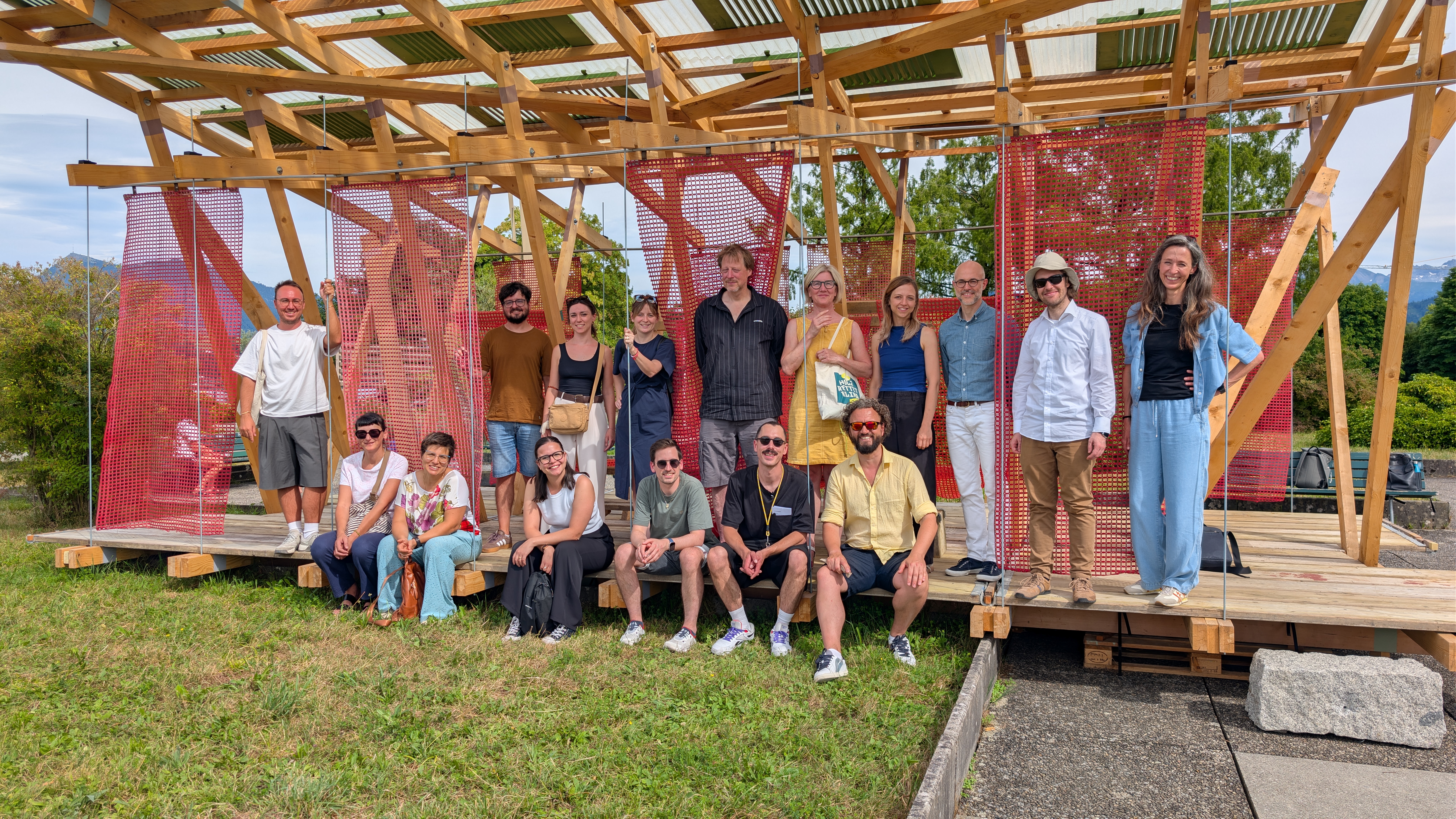 Das Bild zeigt eine Gruppe von Menschen, die vor einer offenen Holzkonstruktion mit roten Netzelementen für ein gemeinsames Foto posiert. Einige stehen, andere sitzen auf einer Holzplattform; im Hintergrund sind Bäume, Berge und ein teilweise bewölkter Himmel zu sehen.