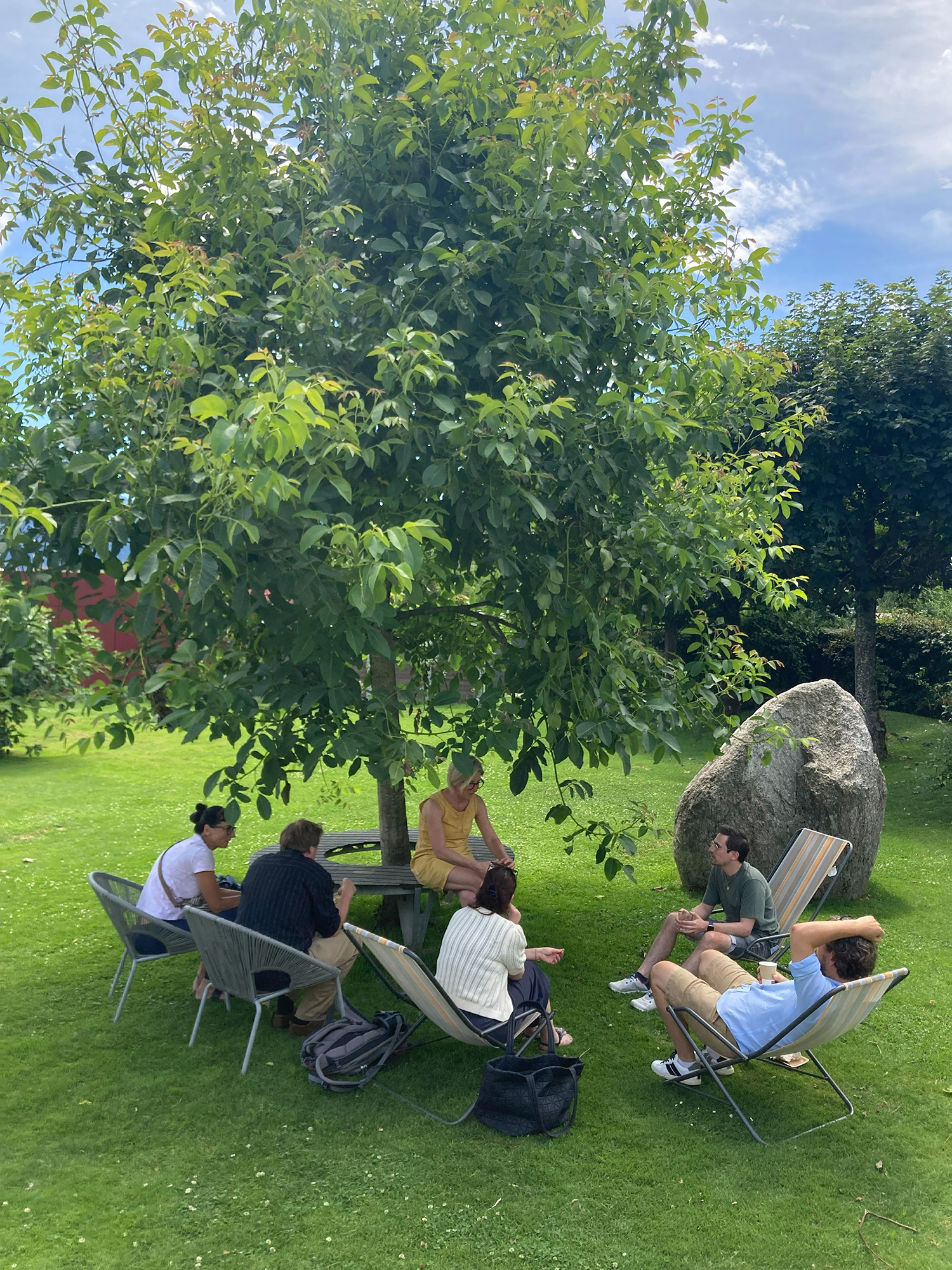 Das Bild zeigt mehrere Personen, die auf einer grünen Wiese unter einem Baum in Liegestühlen und an einem Tisch zusammensitzen. Rechts liegt ein großer Findling, im Hintergrund sind weitere Bäume zu sehen.
