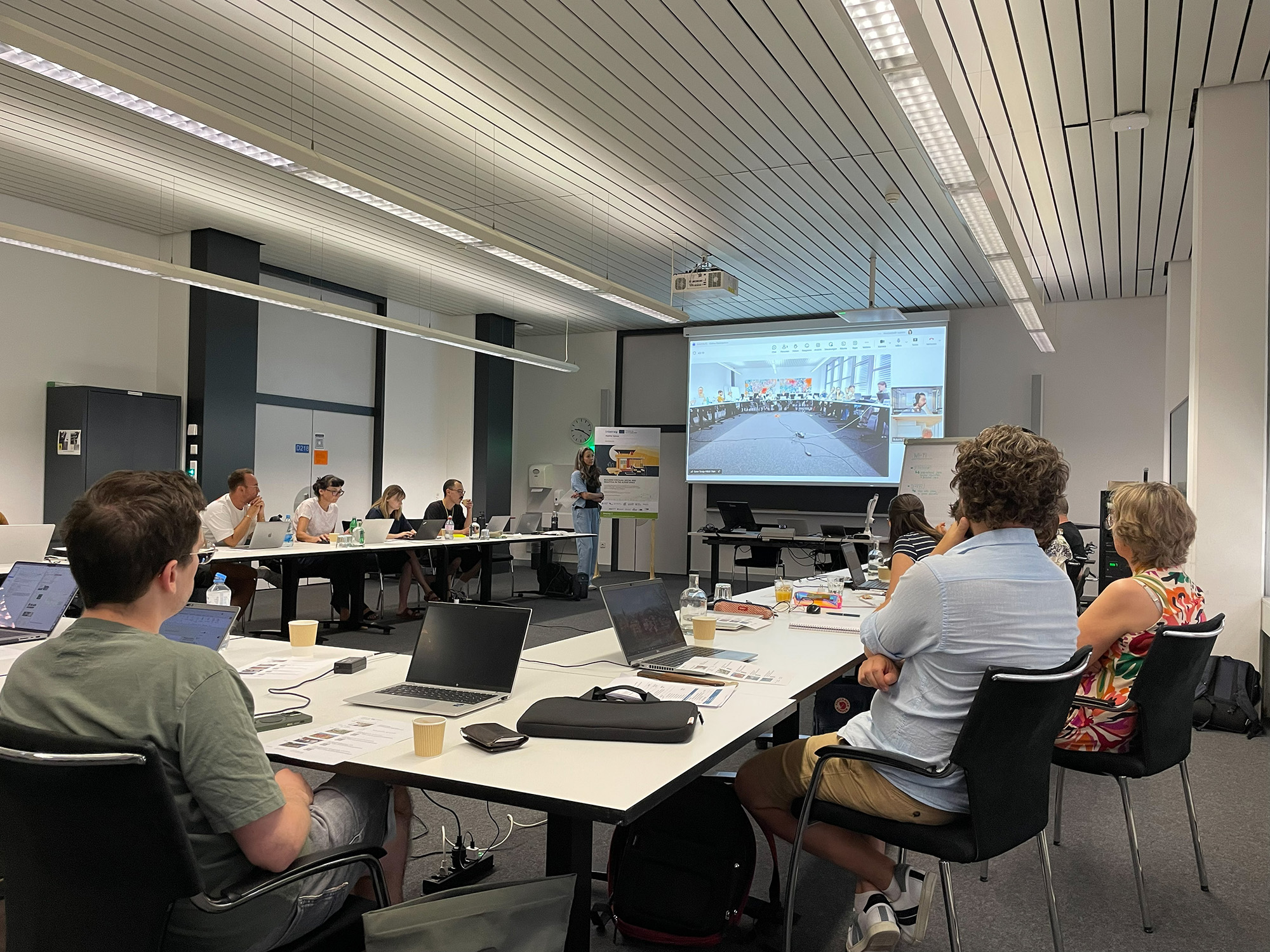 Das Bild zeigt eine Gruppe von Personen in einem Seminarraum mit Laptops an Tischen. Eine Präsentation wird auf eine große Leinwand projiziert.