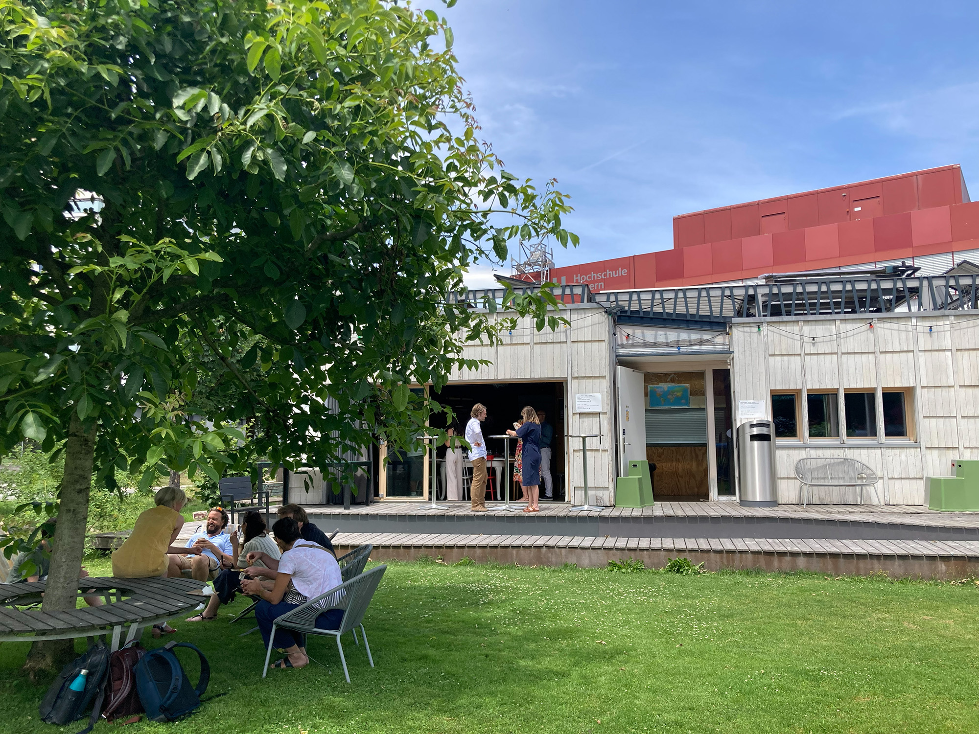 Das Bild zeigt ein helles Holzgebäude auf einem begrünten Gelände der Hochschule Luzern. Im Vordergrund sitzen mehrere Personen an Tischen und unterhalten sich.