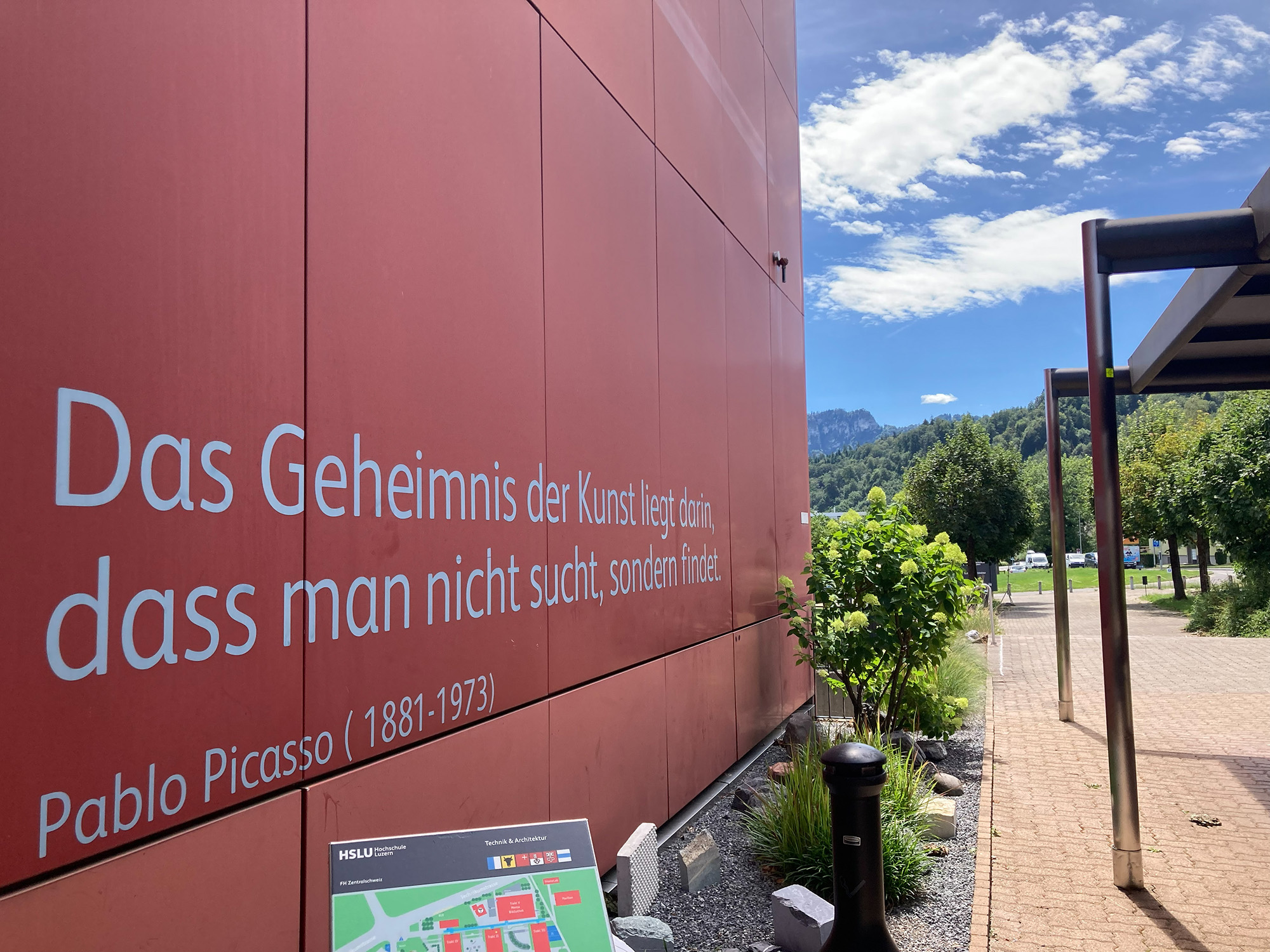 Das Bild zeigt eine rote Gebäudewand mit einem großen Zitat von Pablo Picasso. Daneben führen ein gepflasterter Weg und eine überdachte Konstruktion durch eine begrünte Außenanlage.