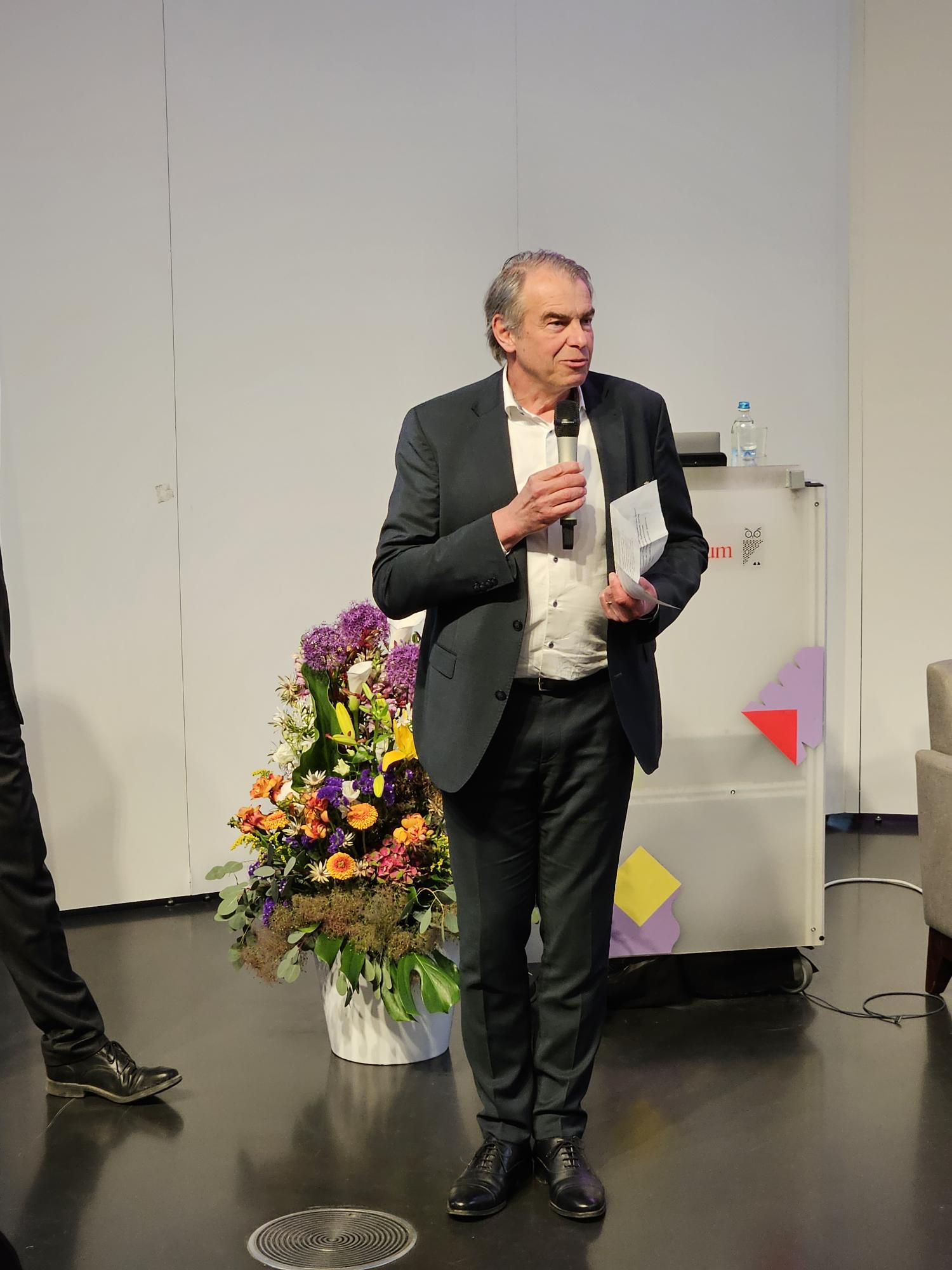 Markus Eder, Ministerialdirigent im Bayerischen Staatsministerium für Wirtschaft, Landes-entwicklung und Energie, spricht in ein Mikrofon, während hinter ihm ein Pult und ein Blu-menstrauß in einer Vase zu sehen sind.