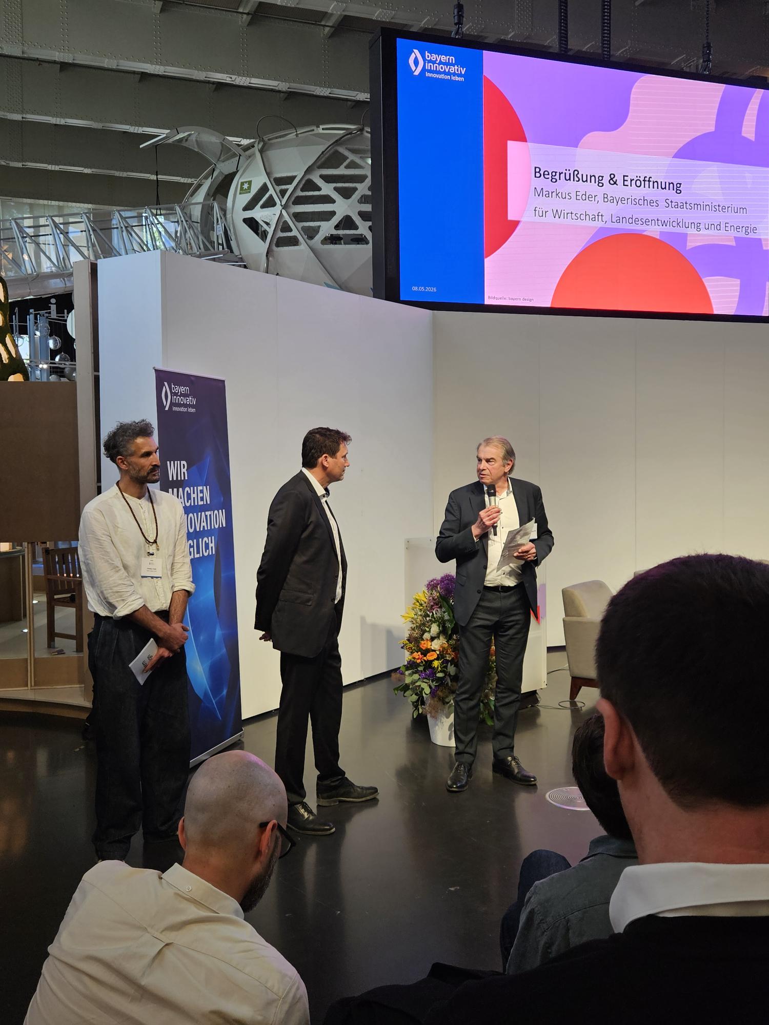 Begrüßung & Eröffnung: Markus Eder, Ministerialdirigent, Bayerisches Staatsministerium für Wirtschaft, Landesentwicklung und Energie, spricht vor Publikum in ein Mikrofon, während Robert Bartl, Mitglied der Geschäftsleitung, Bayern Innovativ GmbH und Christian Fayek, Leitung Projekte, bayern design GmbH, neben ihm stehen.