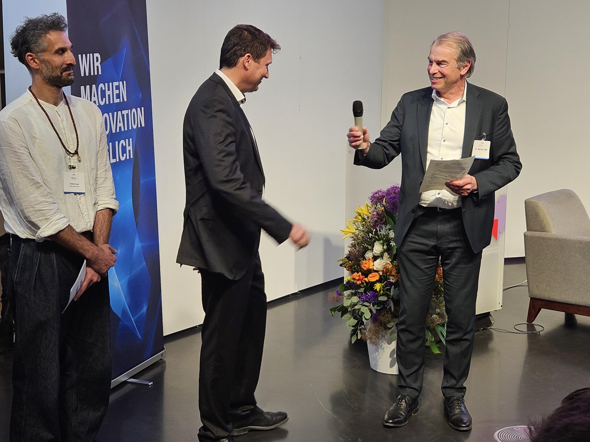 Markus Eder, Ministerialdirigent, Bayerisches Staatsministerium für Wirtschaft, Landesent-wicklung und Energie, überreicht Robert Bartl, Mitglied der Geschäftsleitung, Bayern Innova-tiv GmbH, das Mikrofon, während Christian Fayek, Leitung Projekte, bayern design GmbH, daneben steht.