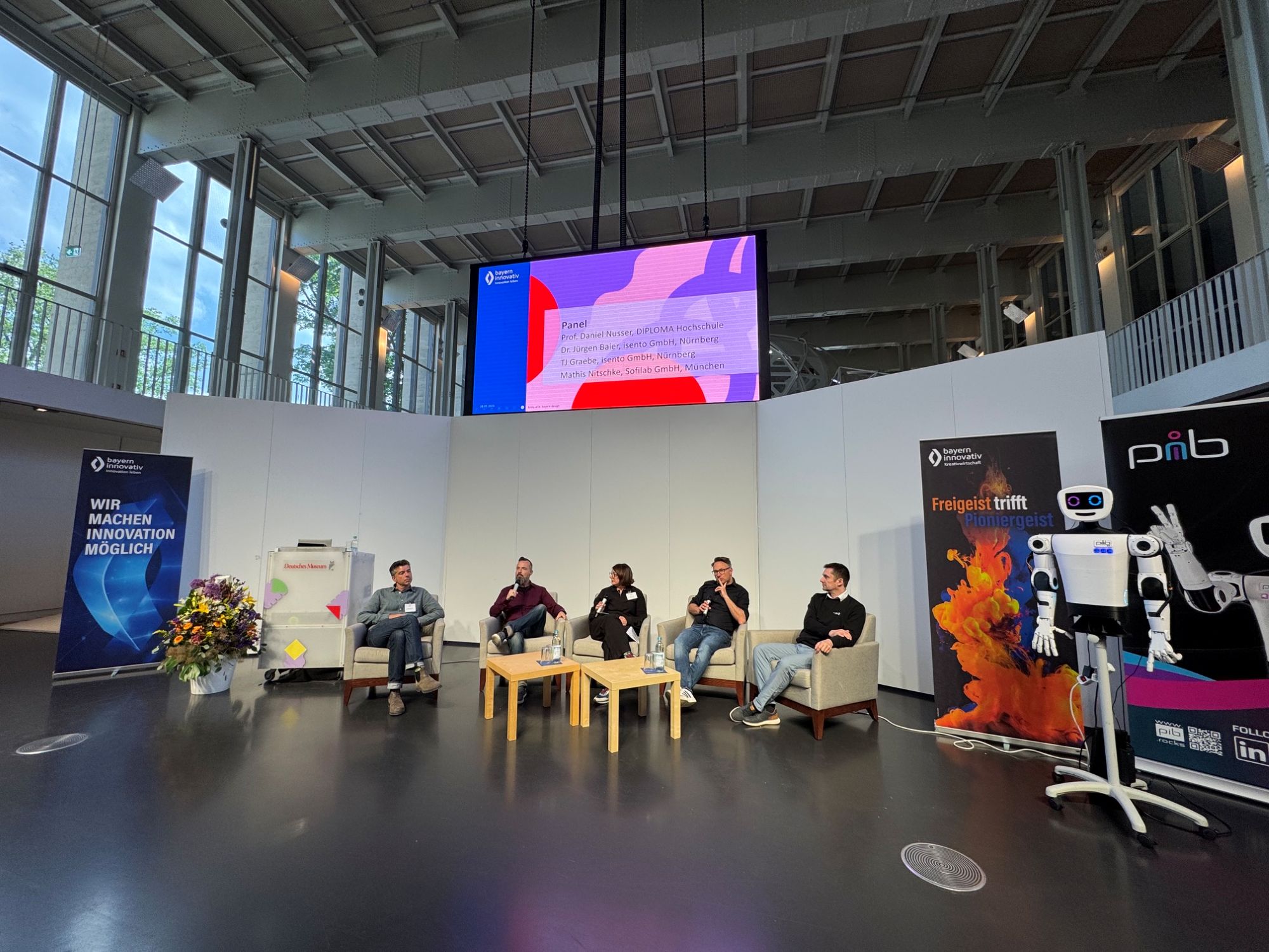 Panel: Mathis Nitschke, Komponist, Technologe und Unternehmer bei der Sofilab GmbH in München, TJ Gräbe, Problemlöser bei der isento GmbH, Prof. Daniel Nusser, Professor und Studiengangsleitung für Prozess- und Produktdesign an der DIPLOMA Hochschule, sowie Dr. Jürgen Baier, Geschäftsführer der isento GmbH, sitzen vorne in Sesseln nebeneinander und diskutieren vor Publikum. Moderiert wird die Runde von Johanna Lison, Projektmanagerin CreativeTechHUB, Bayern Innovativ GmbH. Daneben steht der pib, (printable intelligent bot und ist ein 3D-druckbarer humanoider Roboter).