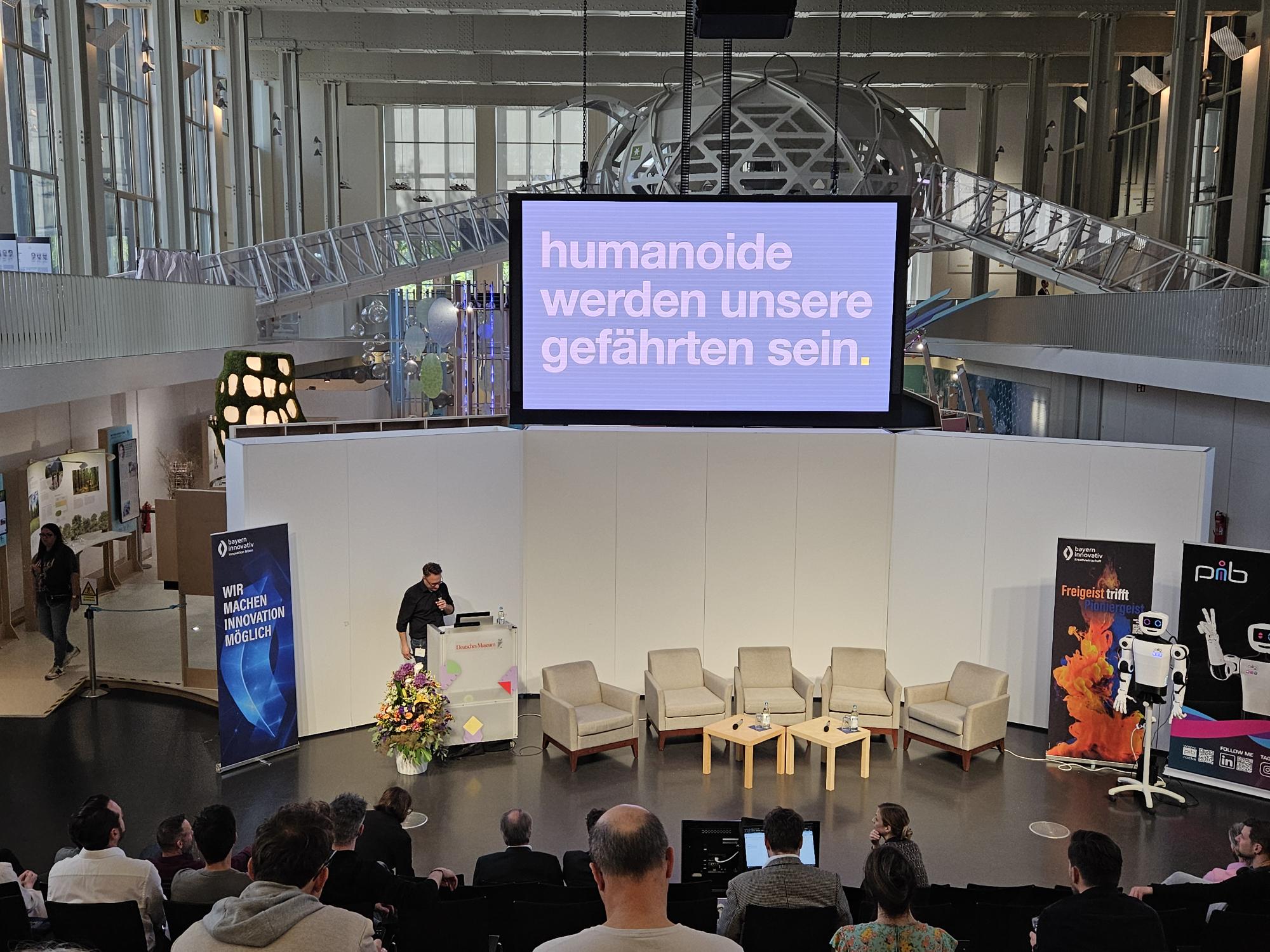 Prof. Daniel Nusser, Professor und Studiengangsleiter für Prozess- und Produktdesign, DIP-LOMA Hochschule, spricht am Pult zum Publikum, während auf dem Bildschirm der Satz „Humanoide werden unsere Gefährten sein“ zu sehen ist.