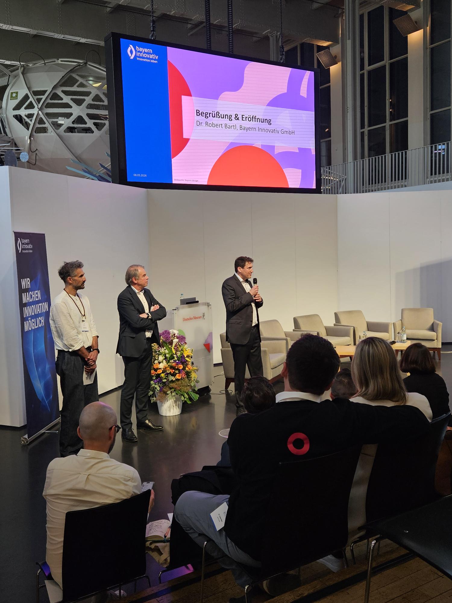Begrüßung & Eröffnung: Robert Bartl, Mitglied der Geschäftsleitung, Bayern Innovativ GmbH, spricht vor Publikum in ein Mikrofon, während Markus Eder, Ministerialdirigent, Bayerisches Staatsministerium für Wirtschaft, Landesentwicklung und Energie und Christian Fayek, Leitung Projekte, bayern design GmbH, neben ihm stehen.