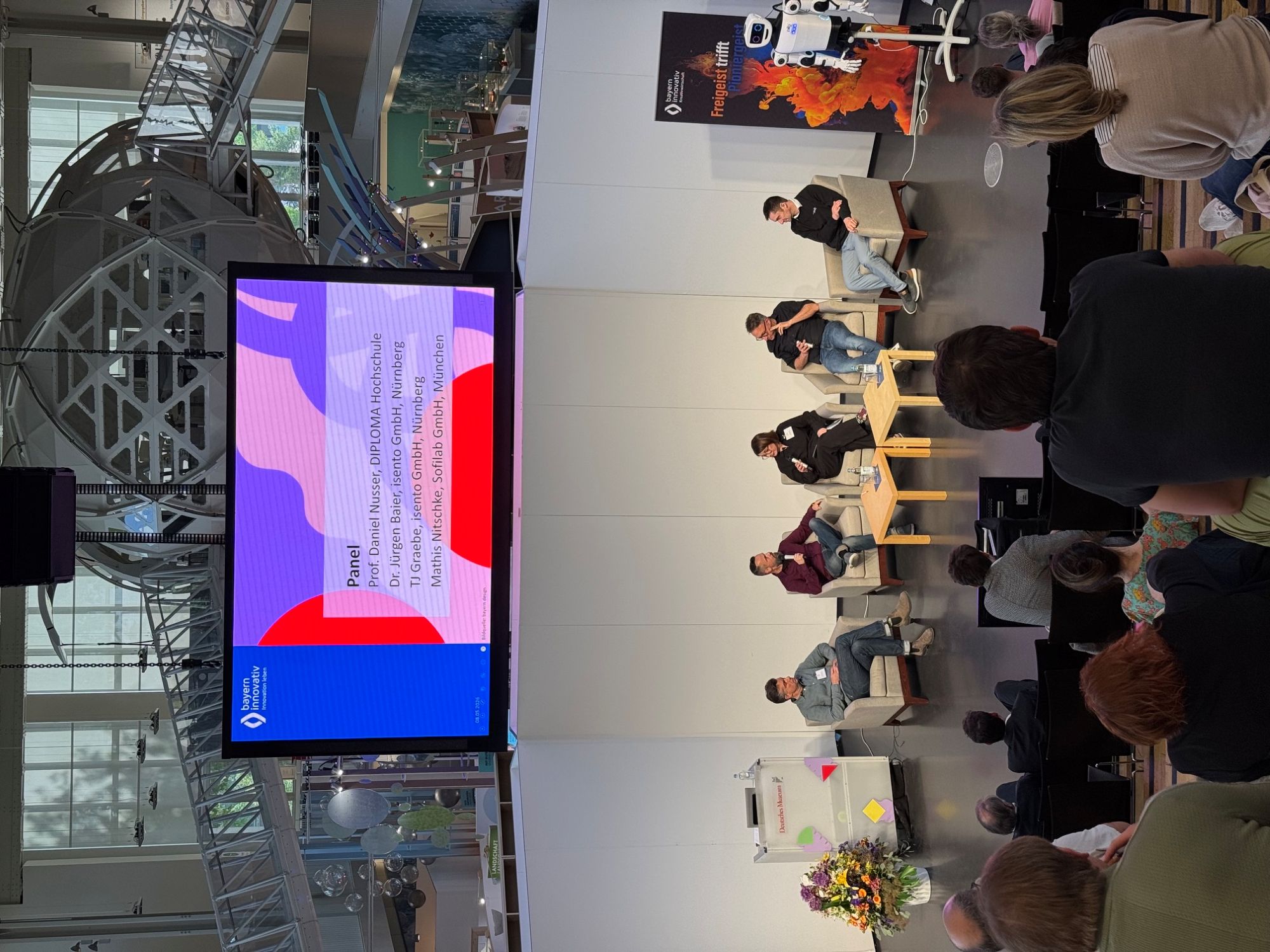 Panel: Mathis Nitschke, Komponist, Technologe und Unternehmer bei der Sofilab GmbH in München, TJ Gräbe, Problemlöser bei der isento GmbH, Prof. Daniel Nusser, Professor und Studiengangsleitung für Prozess- und Produktdesign an der DIPLOMA Hochschule, sowie Dr. Jürgen Baier, Geschäftsführer der isento GmbH, sitzen vorne in Sesseln nebeneinander und diskutieren vor Publikum. Moderiert wird die Runde von Johanna Lison, Projektmanagerin CreativeTechHUB, Bayern Innovativ GmbH.