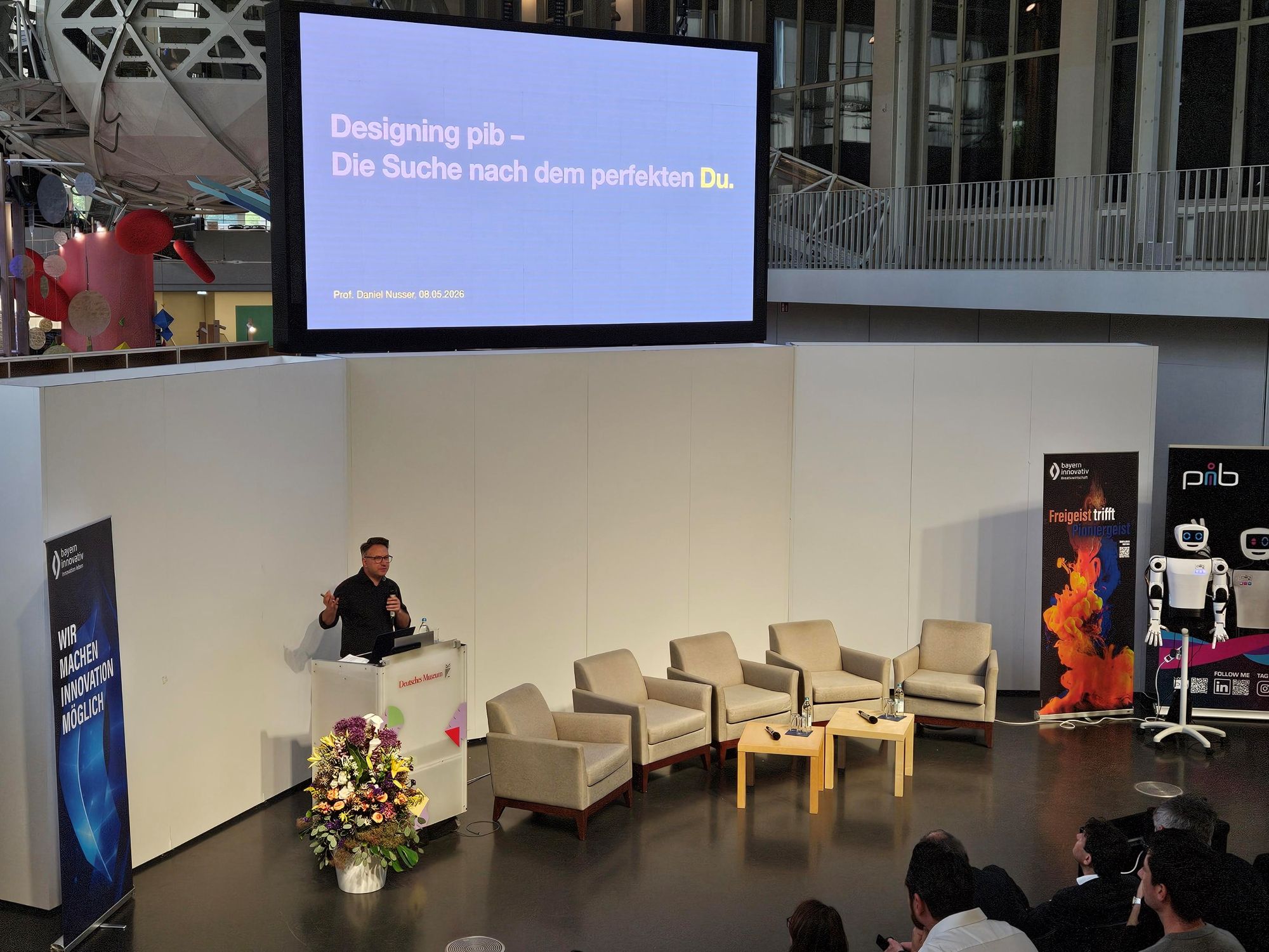 Designing PiB – Die Suche nach dem perfekten Du: Prof. Daniel Nusser, Professor und Studi-engangsleitung für Prozess- und Produktdesign, DIPLOMA Hochschule, ist zu sehen, wie er am Pult ins Mikrofon vor Publikum spricht.