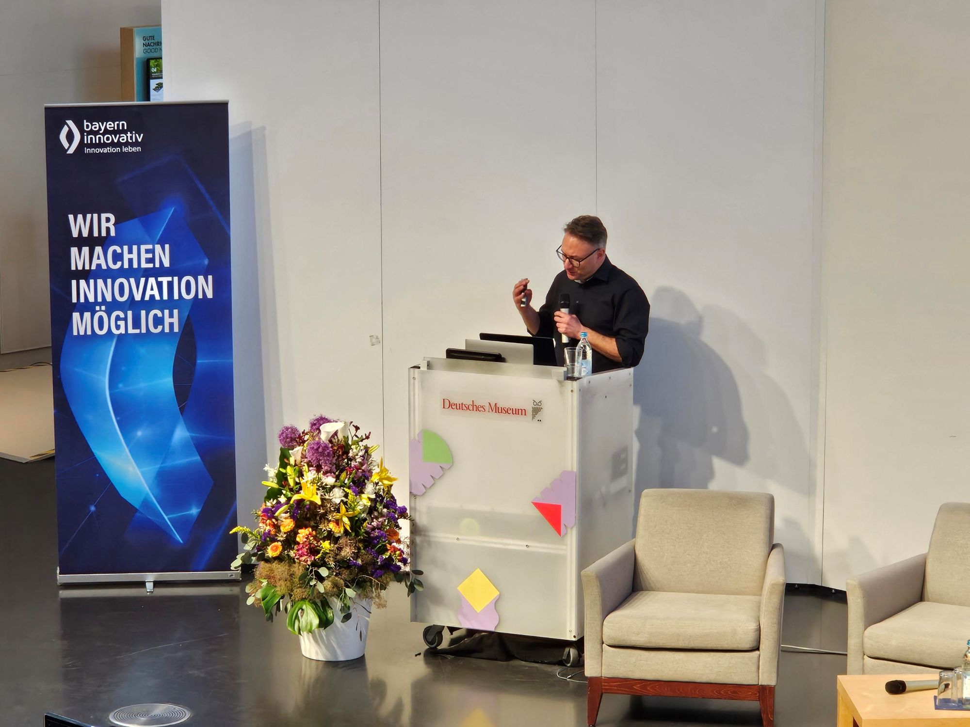Prof. Daniel Nusser, Professor und Studiengangsleitung für Prozess- und Produktdesign, DIP-LOMA Hochschule, ist zu sehen, wie er am Pult ins Mikrofon spricht, während vor ihm eine Vase mit Blumen und ein Roll-up von Bayern Innovativ zu sehen sind.