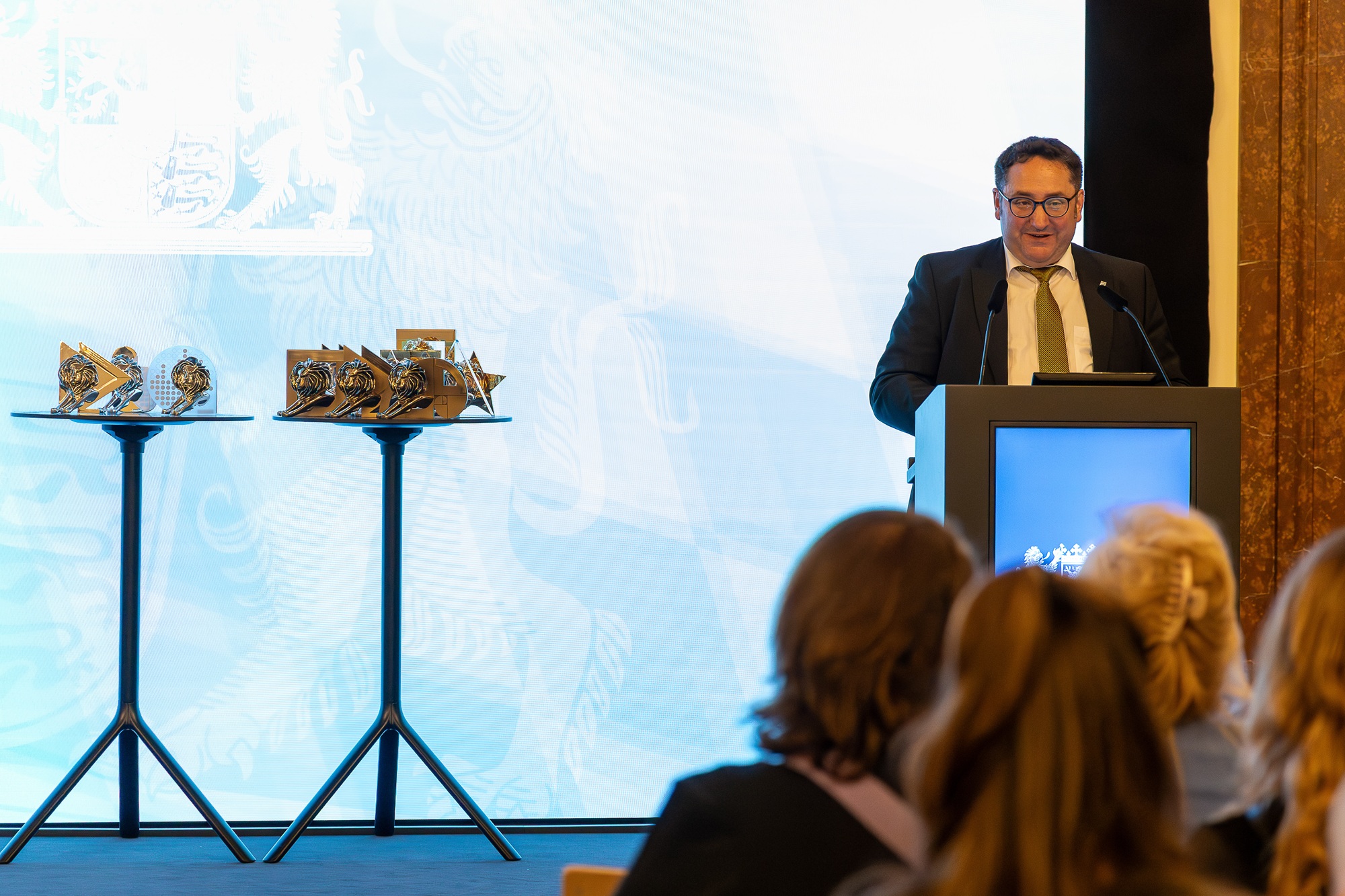 Tobias Gotthardt, Staatssekretär im Bayerischen Staatsministerium für Wirtschaft, Landesentwicklung und Energie, steht an einem Rednerpult und spricht vor Publikum. Neben ihm befinden sich zwei Stehtische mit goldfarbenen Trophäen.