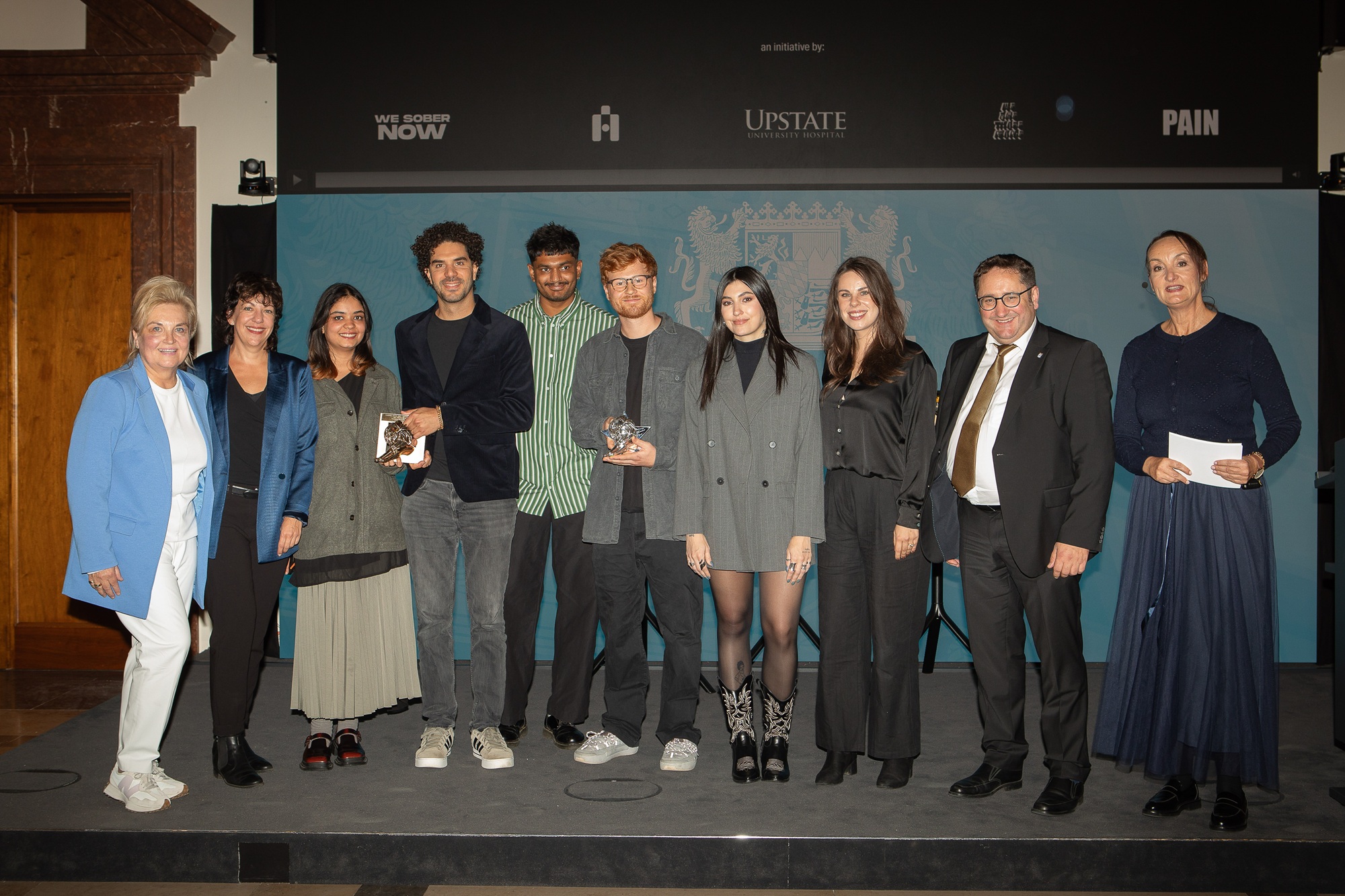 Tobias Gotthardt, Staatssekretär im Bayerischen Staatsministerium für Wirtschaft, Landesentwicklung und Energie, und Petra Lorenz, Geschäftsführerin des CommClubs Bayern e.V., stehen gemeinsam mit bayerischen Gewinnern und Gewinnerinnen des diesjährigen Cannes Lions auf einer Bühne.