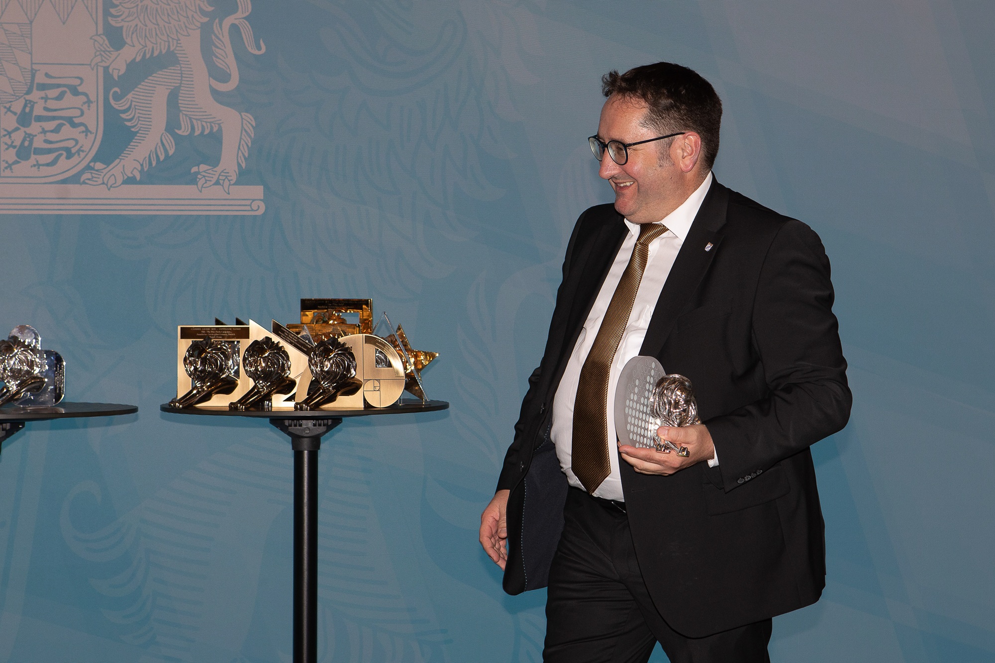 Tobias Gotthardt, Staatssekretär im Bayerischen Staatsministerium für Wirtschaft, Landesentwicklung und Energie, steht auf einer Bühne und hält eine silberne Trophäe in der Hand. Auf den Tischen neben ihm sind weitere Trophäen zu sehen.