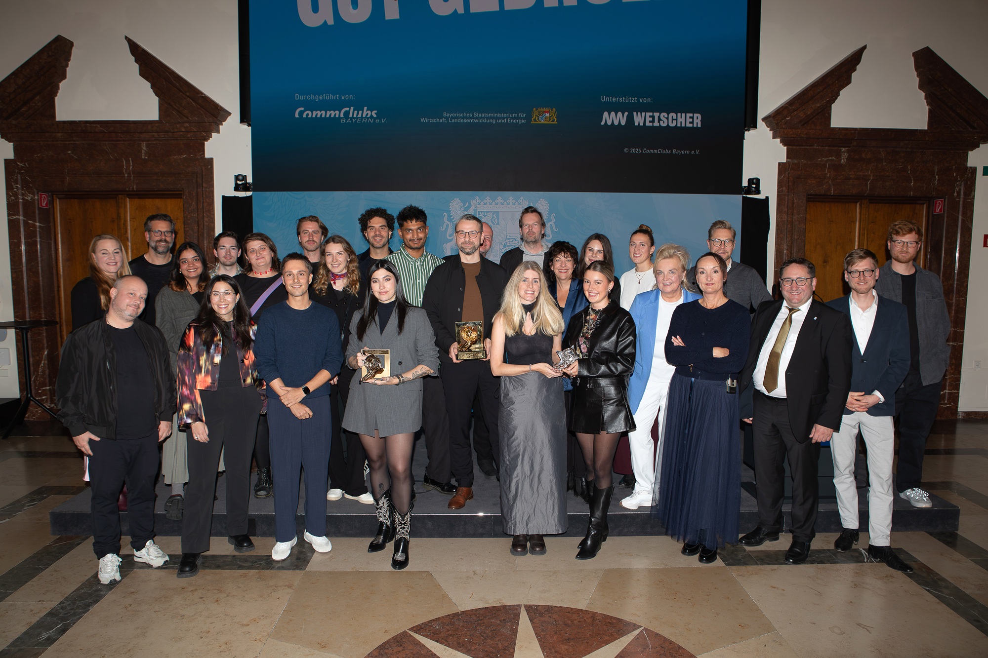 Tobias Gotthardt, Staatssekretär im Bayerischen Staatsministerium für Wirtschaft, Landesentwicklung und Energie, und Petra Lorenz, Geschäftsführerin des CommClubs Bayern e.V., stehen gemeinsam mit bayerischen Gewinnern und Gewinnerinnen des diesjährigen Cannes Lions auf einer Bühne.