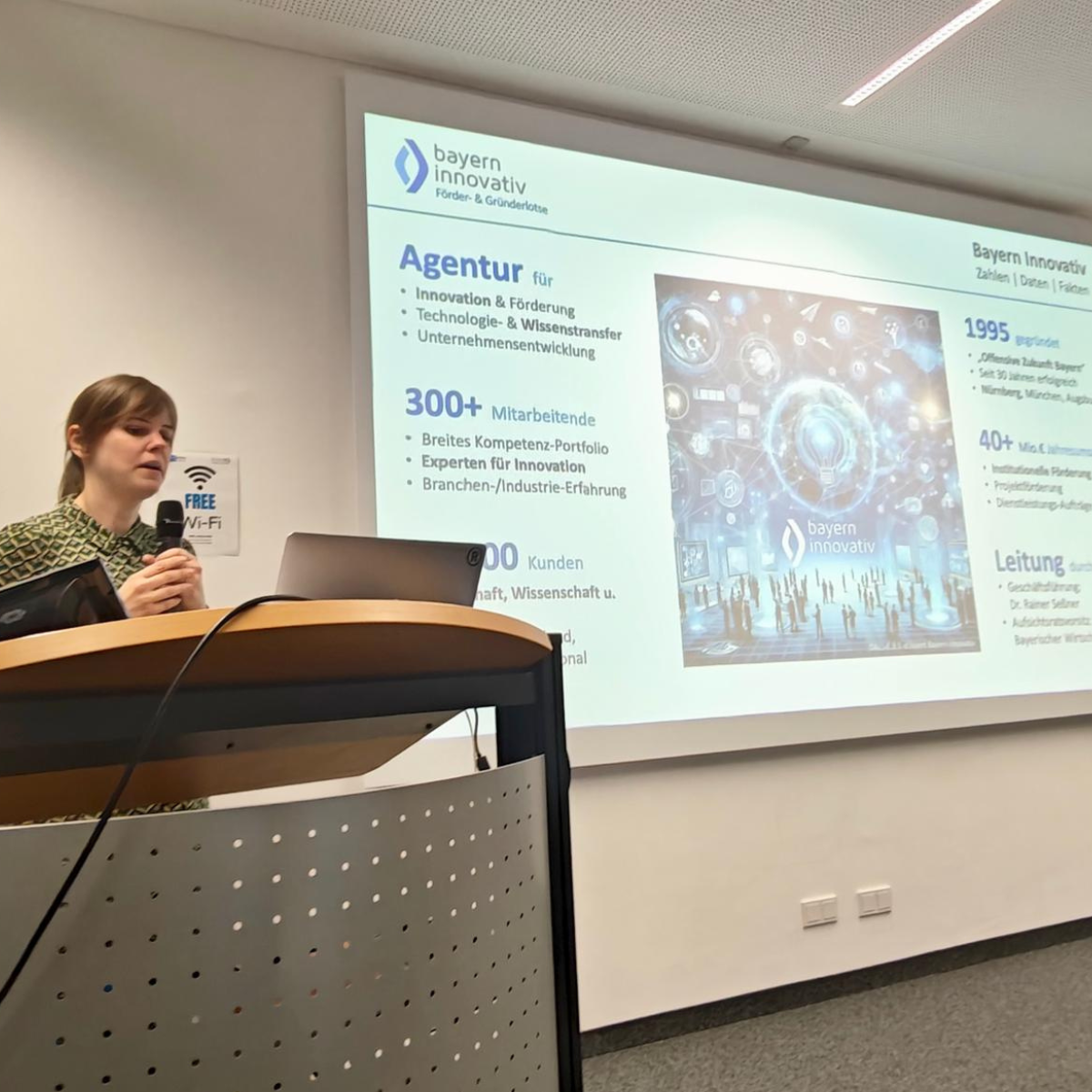 Eine Frau steht hinter einem Podium und spricht. Im Hintergrund sieht man die Folien einer Powerpointpräsentation