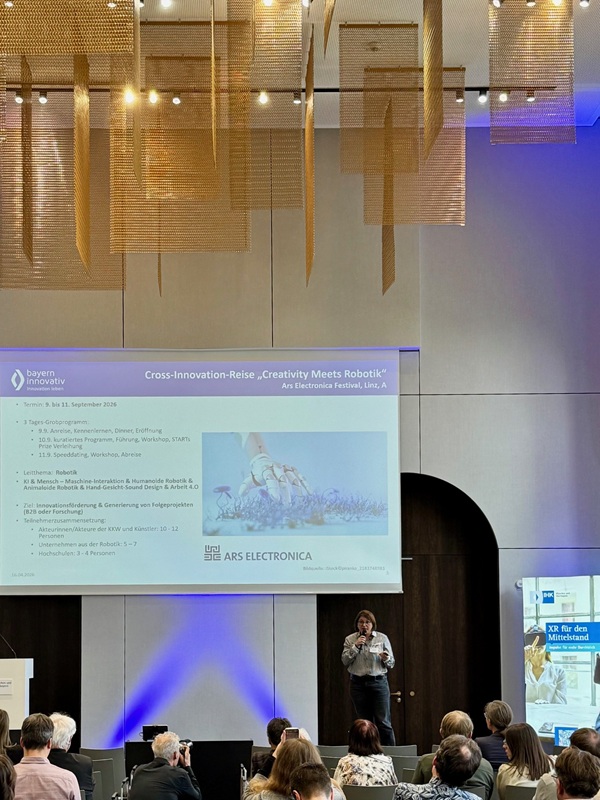 Im Konferenzraum der Veranstaltung ‚XR für den Mittelstand‘ spricht Projektmana-gerin Johanna Lison (Bayern Innovativ GmbH) vor Publikum auf der Bühne mit Mik-rofon, während im Hintergrund eine Präsentationsfolie gezeigt wird.