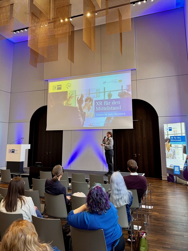 Im Konferenzraum der Veranstaltung ‚XR für den Mittelstand‘ spricht Projektmana-gerin Johanna Lison (Bayern Innovativ GmbH) vor Publikum auf der Bühne mit Mik-rofon, während im Hintergrund eine Präsentationsfolie gezeigt wird.