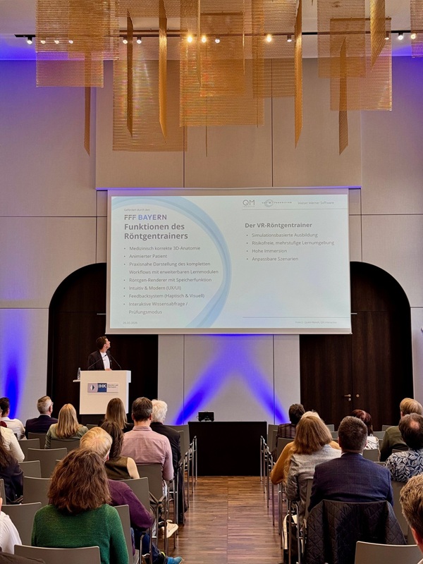 Im Konferenzraum spricht Geschäftsführer Quirin Münch (CAT Production) bei der Veranstaltung ‚XR für den Mittelstand‘ vor Publikum an einem Sprecherpult, wäh-rend er auf eine Präsentationsfolie schaut. 