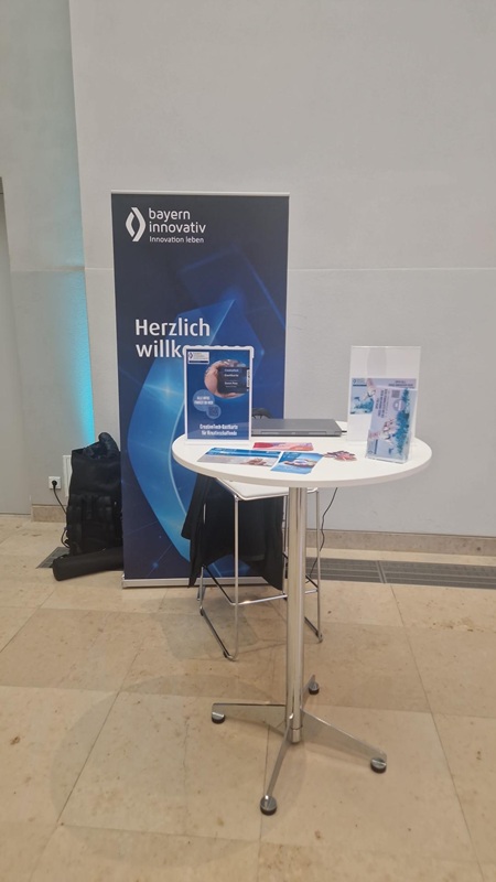In einem Innenraum steht ein Tisch, auf dem Flyer liegen, dahinter ist ein Roll-up-Banner von Bayern Innovativ zu sehen.