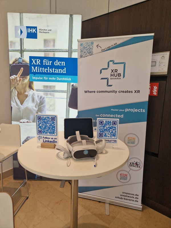 Auf einem runden Tisch liegen eine VR-Brille und mehrere QR-Codes, dahinter ste-hen zwei Roll-up-Banner mit Informationen zu XR und dem XR Hub Bavaria.