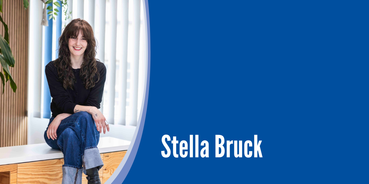 Frau mit langen, dunklen Haaren sitzt auf einer Holztruhe. Ihre Beine sind überschlagen und ihre Arme liegen locker auf. Sie lächelt aufgeschlossen. 