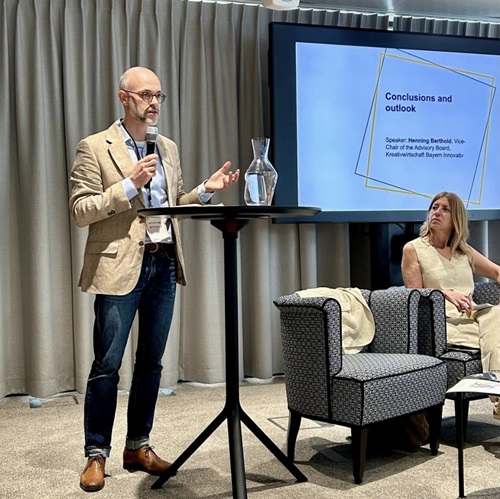 Zu sehen ist Henning Berthold wie er mit einem Mikrofon vor einem Hochtisch stehend zum Publikum redet. Rechts daneben sitzt eine Frau, die ihm gespannt zuhört. Im Hintergrund ist eine eingeblendete Folie zu erkennen, auf der "Coclusions and outlook" zu lesen ist.