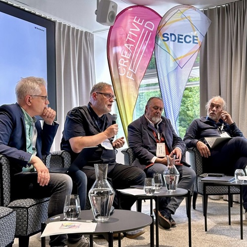 Zu sehen sind vier männliche Personen, die in einer Diskussion zu sein scheinen. Eine Person hält ein Mikrofon während die anderen Personen teils nachdenklich, teils gespannt dem Redenden zuhören. Im Hintergrund sind Beachflags der Creative FED zu sehen. Es handelt sich bei dieser Szene um eine Paneldiskussion. 