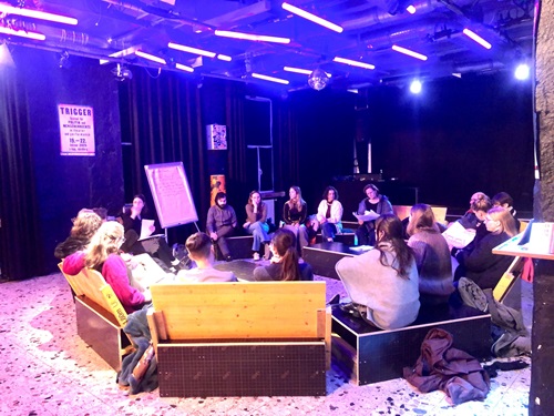 Zu sehen ist eine Gruppe, die in einem Stuhlkreis sitzt. Die Aufnahme ist außerhalb der Sitzgruppe aufgenommen, weswegen die Personen nicht detailliert erkennbar sind. Die Gruppe sitzt in einem Veranstaltungsraum mit violettem/ dunklem Licht. 