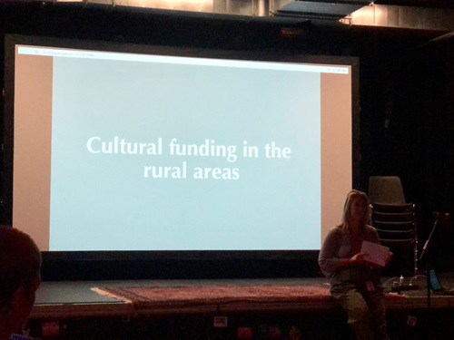 Zu sehen ist eine Bühne, auf der eine Person am rechten Rand sitzt. Darüber ist eine Folie eingeblendet auf der "cultural  funding in the rural areas" zu lesen ist. 