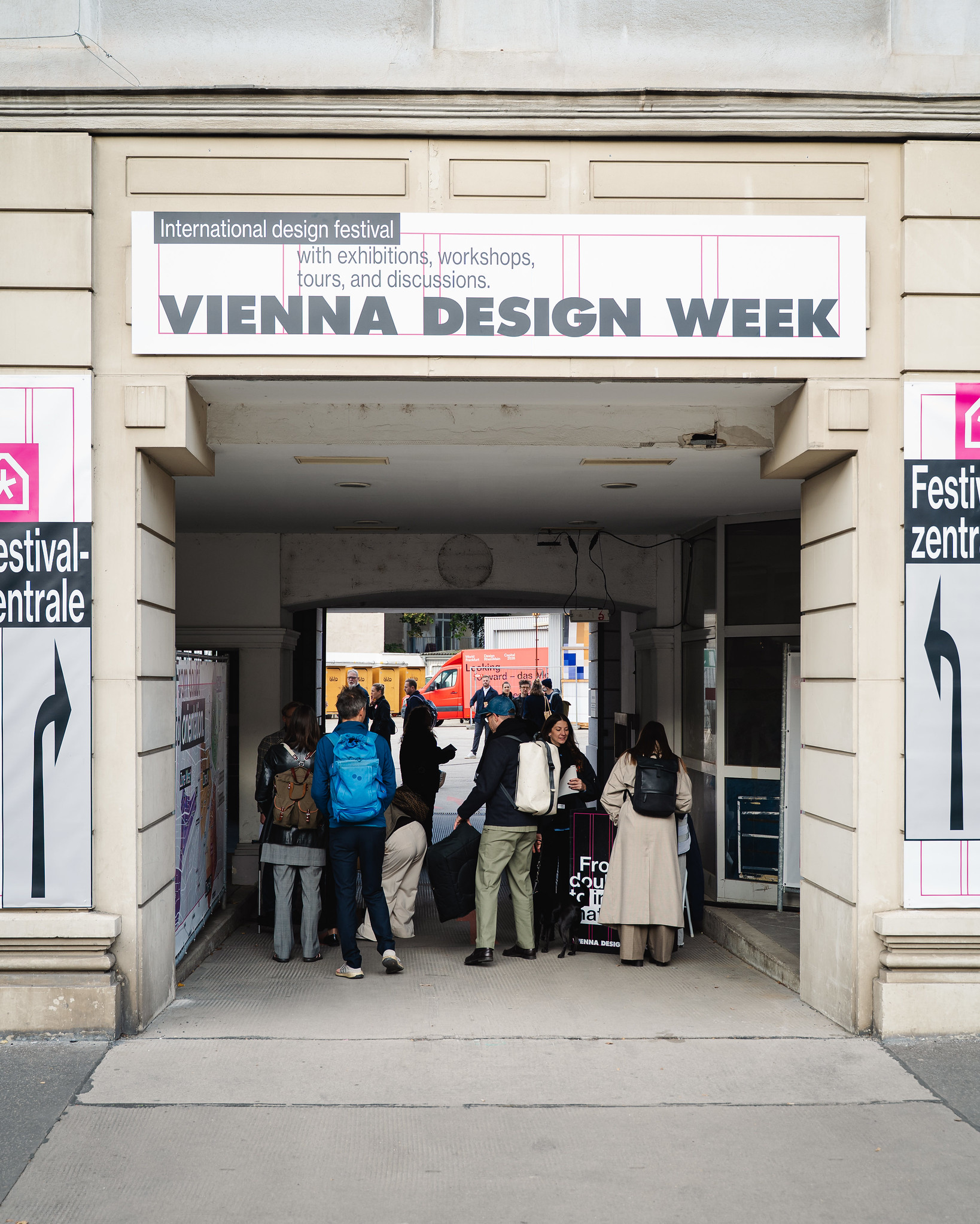 Mehrere Menschen stehen in einem überdachten Durchgang unter einem Schild mit der Aufschrift „Vienna Design Week“. Der Durchgang führt zu einer Straße, wo man im Hintergrund weitere Passanten sieht.