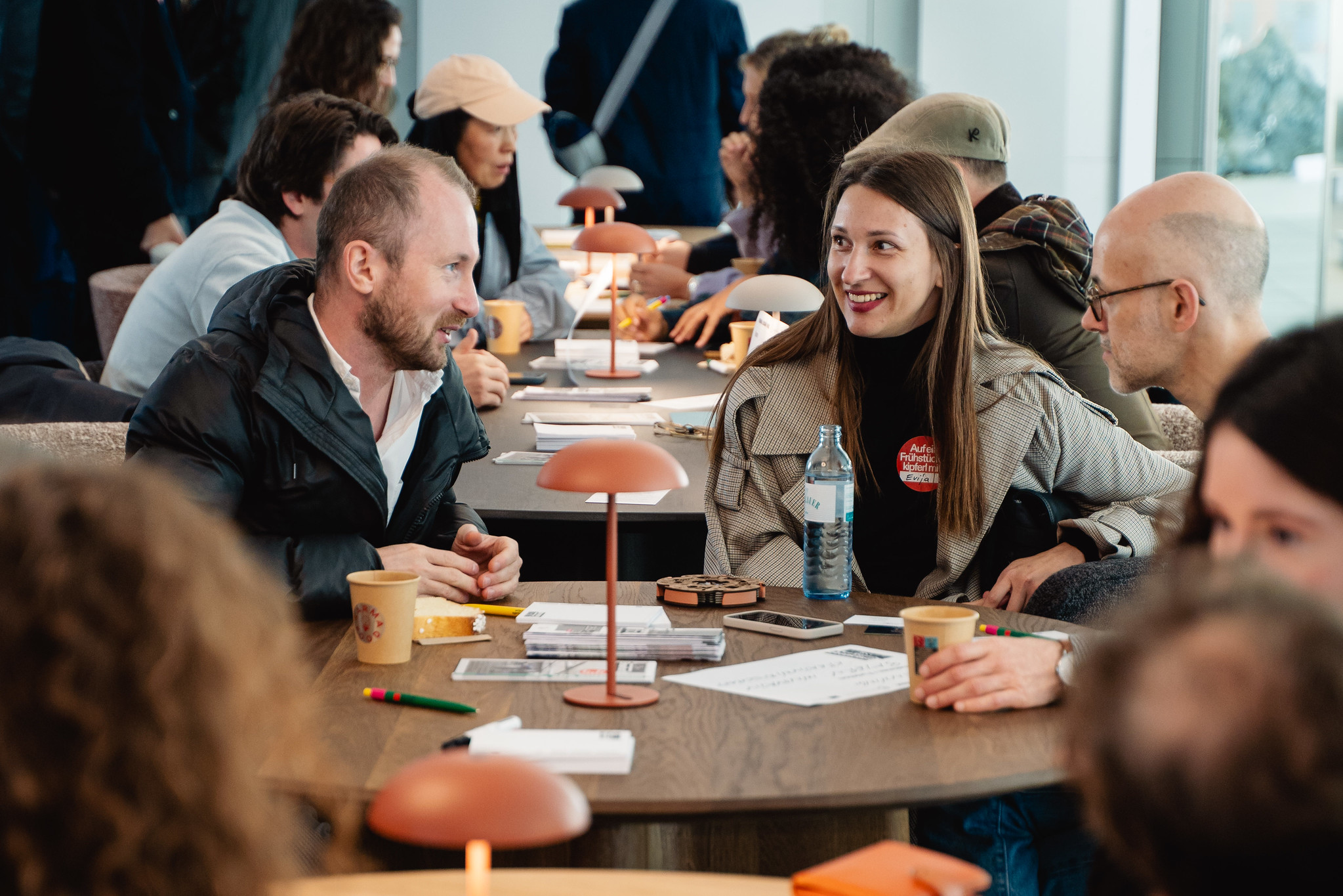 Mehrere Personen sitzen an runden Holztischen und unterhalten sich. Im Vordergrund spricht Henning Berthold, Projektleiter Analyse und Dialog, Innovationsnetzwerk Kreativwirtschaft der Bayern Innovativ GmbH, mit einer Frau und einem weiteren Mann.