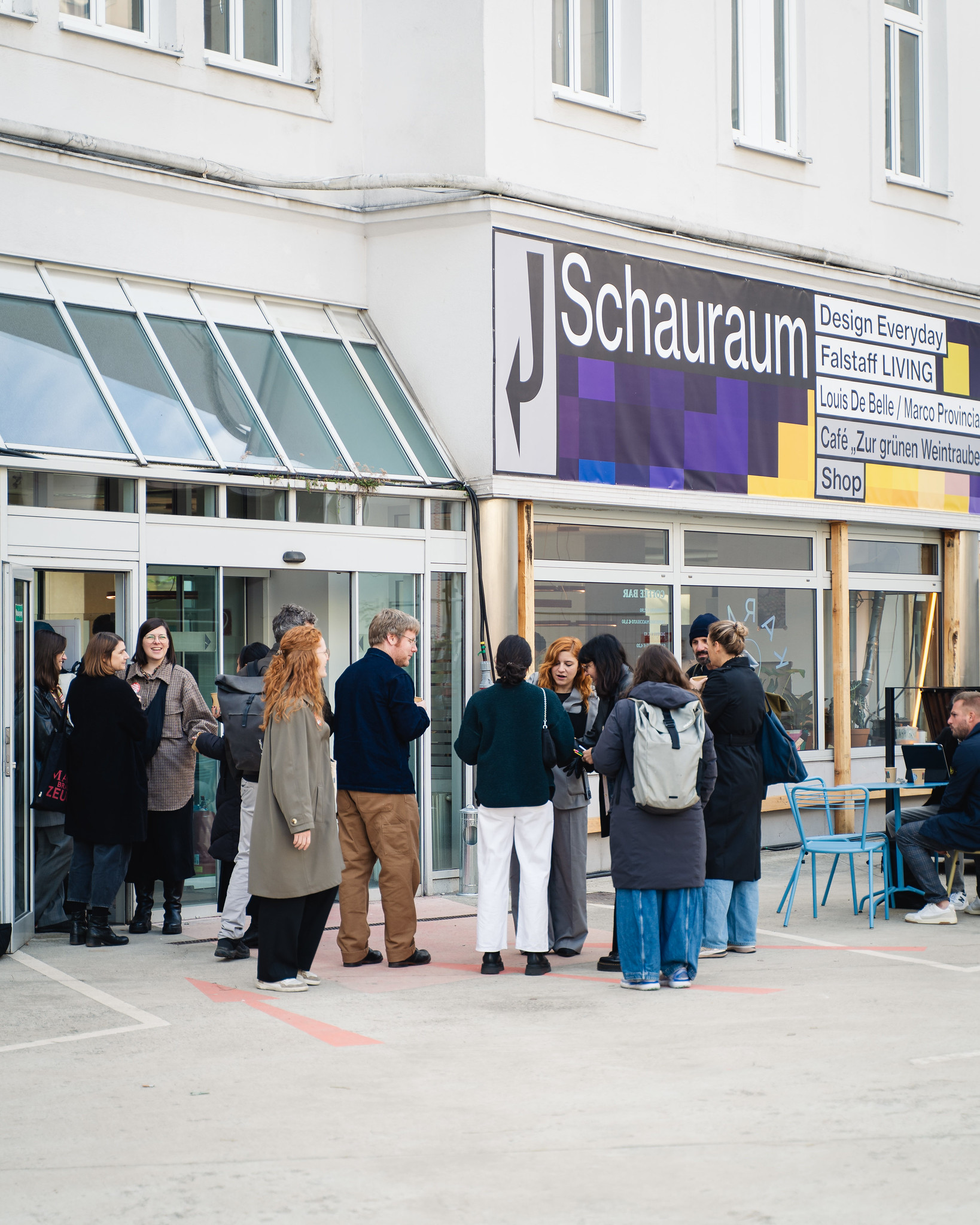 Vor einem Gebäude steht eine Gruppe von Personen, die sich unterhält. Oberhalb und leicht seitlich des Eingangs hängt an der hellen Fassade ein großes, buntes Plakat mit der Aufschrift „Schauraum“.
