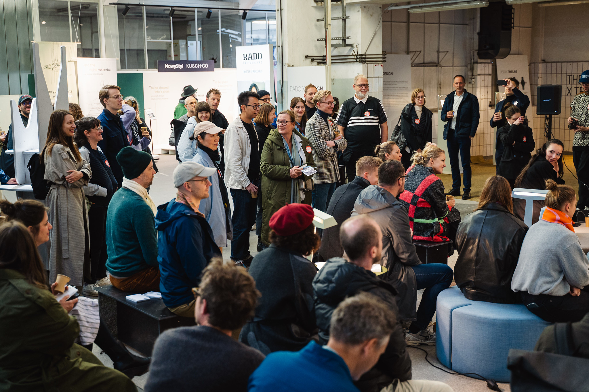 In einem Raum sitzen und stehen einige Personen dicht beieinander und verfolgen eine Veranstaltung. Im Hintergrund sind weiße Ausstellungswände mit Schriftzügen und Logos wie „RADO“, „NowyStyl“ und „KUSCH+CO“ zu sehen.