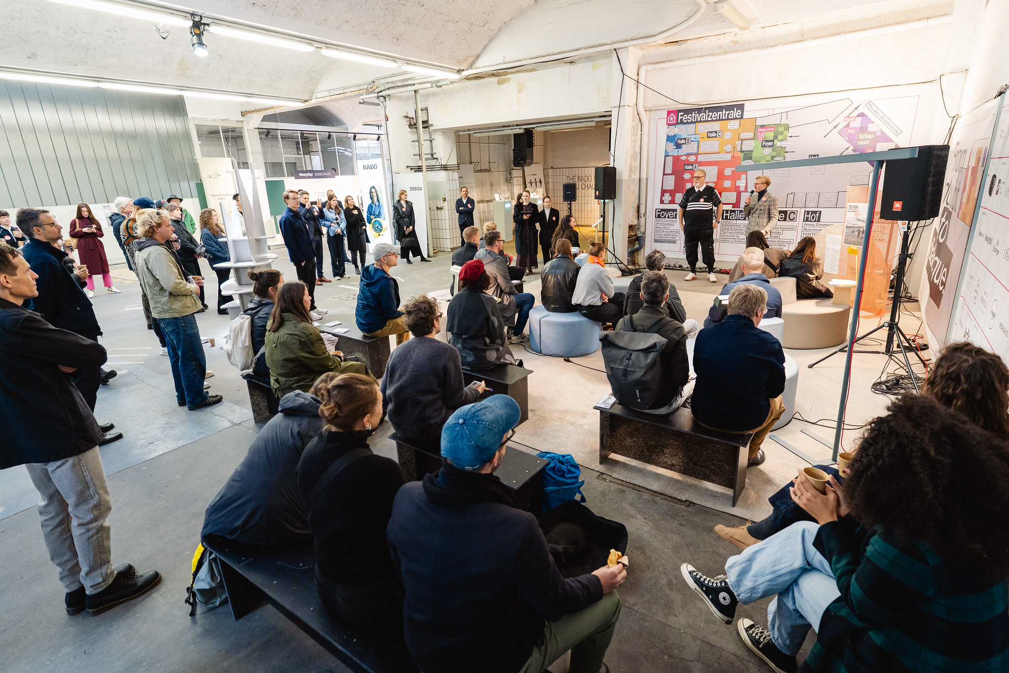 In einem Raum stehen und sitzen mehrere Personen und hören einer Veranstaltung zu. Vorne auf einer kleinen Bühne sprechen zwei Personen mit Mikrofonen. Hinter ihnen sind Infowände mit dem Schriftzug „Festivalzentrale“ und Lageplänen der Räume zu sehen.