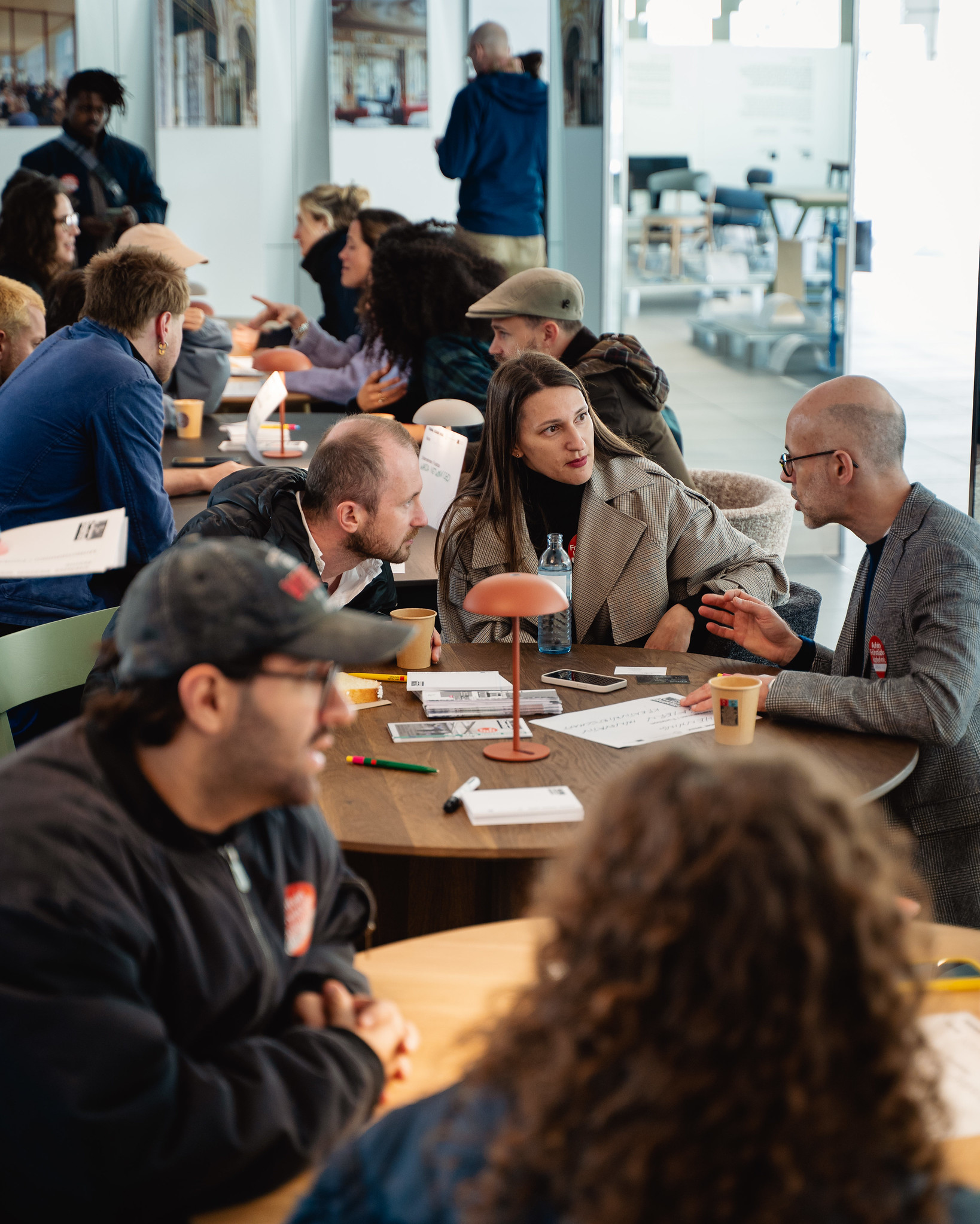 Mehrere Personen sitzen an runden Holztischen und führen Gespräche. Unter anderem spricht Henning Berthold, Projektleiter Analyse und Dialog, Innovationsnetzwerk Kreativwirtschaft der Bayern Innovativ GmbH, mit einer Frau und einem weiteren Mann.