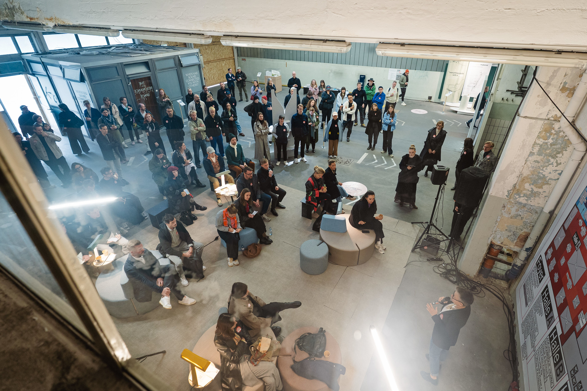 Der Blick von oben zeigt einen Raum, in dem Personen sitzen oder weiter hinten stehen und einer Veranstaltung zuhören. Vorne rechts steht eine Person mit Mikrofon und spricht zu den Zuhörenden. Auf einer Wand ist ein großer Plan mit Beschriftungen wie „Halle A“ und „Halle B“ zu sehen.