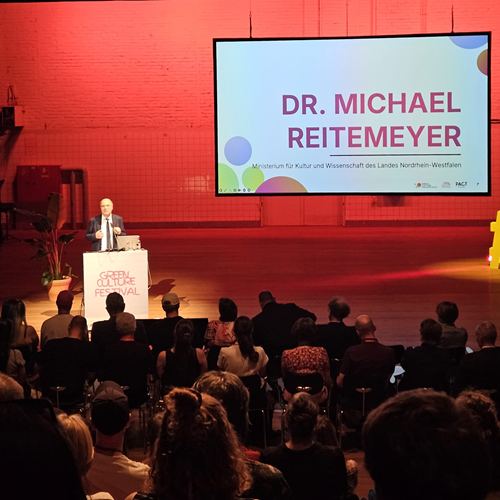 Zu sehen ist eine Bühne mit Rednerpult, hinter der Dr. Michael Reitemeyer eine Rede hält. Die Bühne ist in rotes Licht getaucht. Im unteren Teil des Bildes ist ein Teil des Publikums zu sehen.