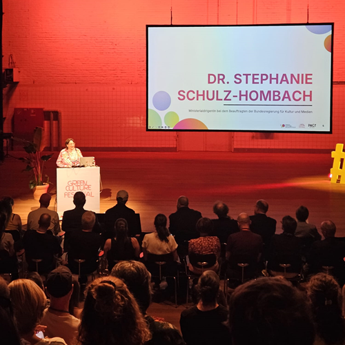 Zu sehen ist eine Bühne mit Rednerpult, hinter der Dr. Stephanie Schulz-Hombach eine Rede hält. Die Bühne ist in rotes Licht getaucht. Im unteren Teil des Bildes ist ein Teil des Publikums zu sehen.