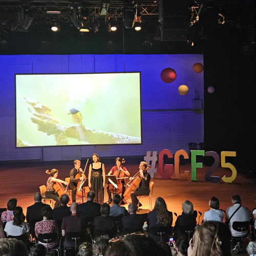 Zu sehen ist eine Bühne auf der ein Streichquartett sowie eine Sängerin mittig platziert sind. Im Hintergrund sind Visualisierungen eines Vogels zu sehen. Im Vordergrund ein Teil des Publikums. 