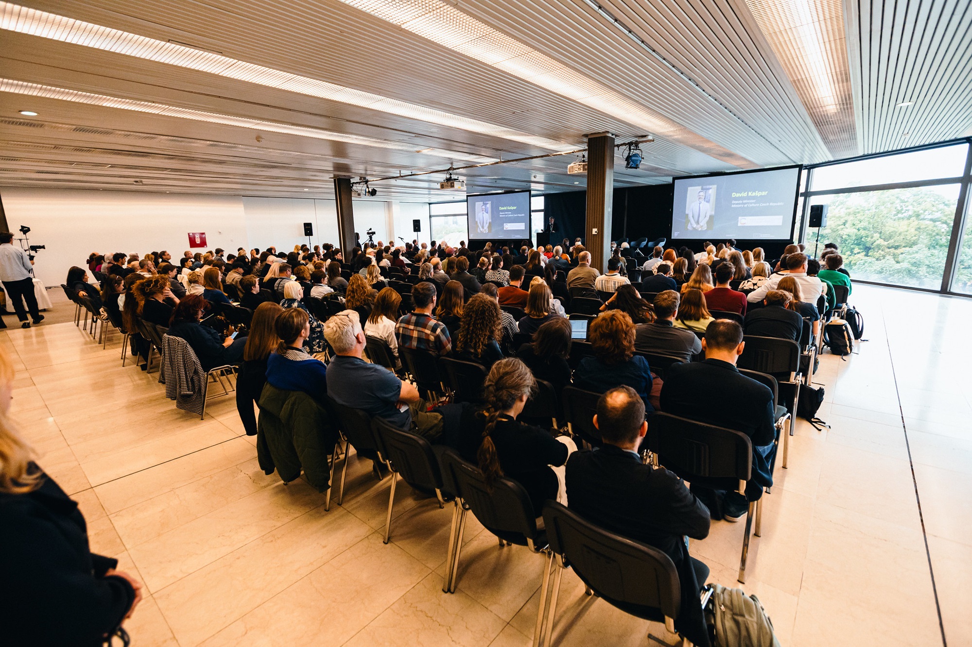 In einem großen Konferenzraum sitzen einige Personen auf Stuhlreihen und verfolgen eine Präsentation. Vorne steht eine Person auf einer Bühne, daneben sind zwei große Leinwände mit einer projizierten Folie zu sehen.