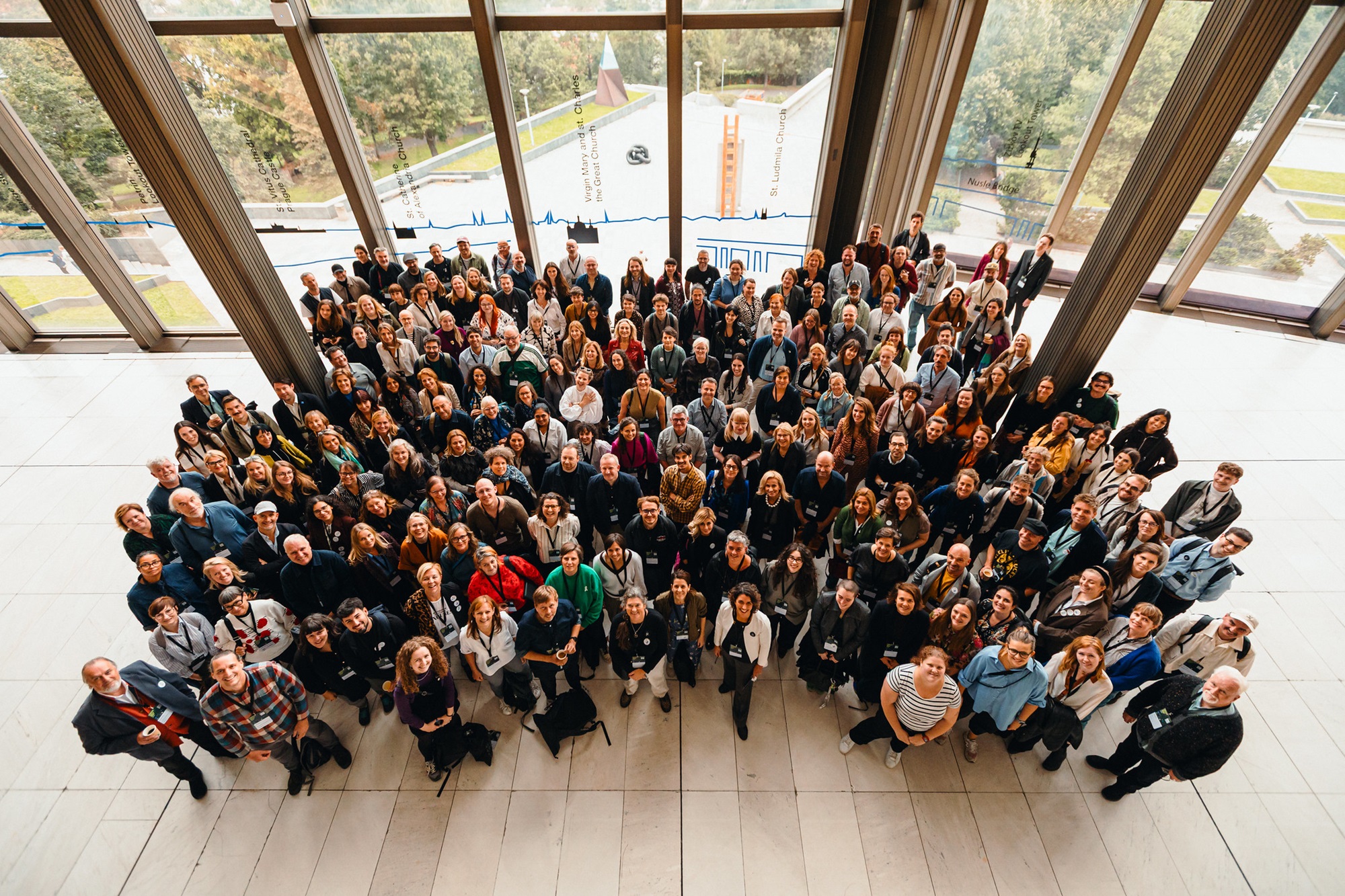 Ein von oben aufgenommenes Gruppenbild zeigt einige Menschen, die zur Kamera hinaufblicken. Sie sind in mehreren Reihen dicht beieinander angeordnet und lächeln. Durch die großen Fenster im Hintergrund sind Bäume und ein Teil eines Daches zu sehen. 